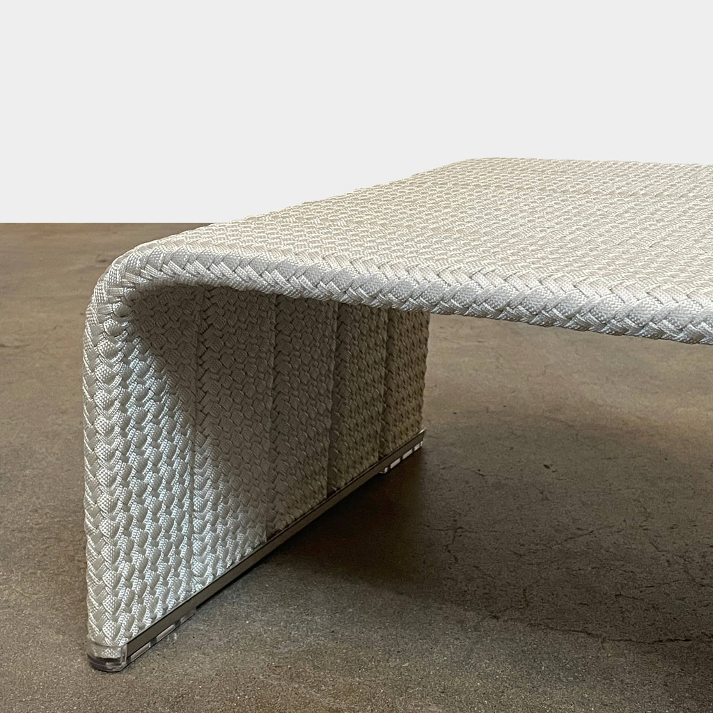 A rectangular woven ottoman with a curved design and a light gray color, this piece showcases superior craftsmanship. The surface texture resembles a braided pattern while the bottom edges feature protective covering caps, making it an ideal match for the Paola Lenti Paola Lenti Frame Outdoor Side Table (ON HOLD).