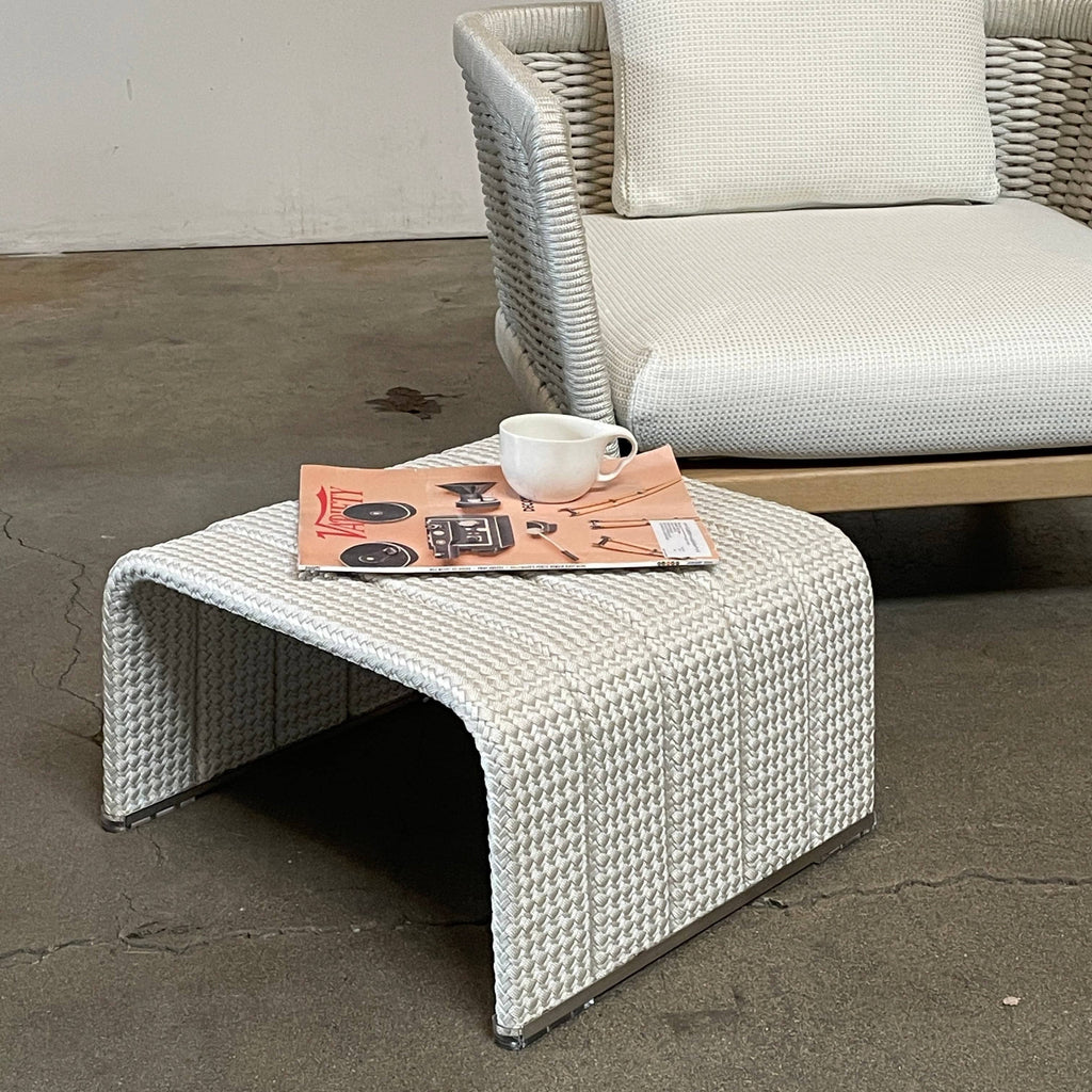 A rectangular woven ottoman with a curved design and a light gray color, this piece showcases superior craftsmanship. The surface texture resembles a braided pattern while the bottom edges feature protective covering caps, making it an ideal match for the Paola Lenti Paola Lenti Frame Outdoor Side Table (ON HOLD).