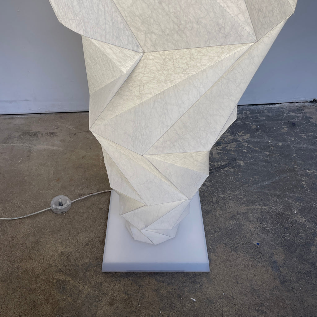 A tall geometric sculpture made of interlocking white panels stands on a simple base, functioning as an Artemide Minomushi floor light in an indoor setting with a concrete floor.