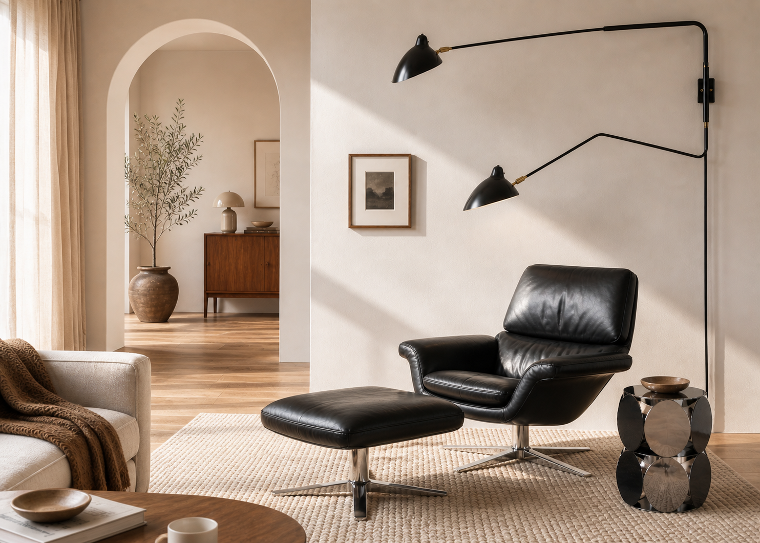 A modern living room with a black leather armchair and ottoman, a wall-mounted lamp, a side table with decor, neutral walls, and natural light from a window.