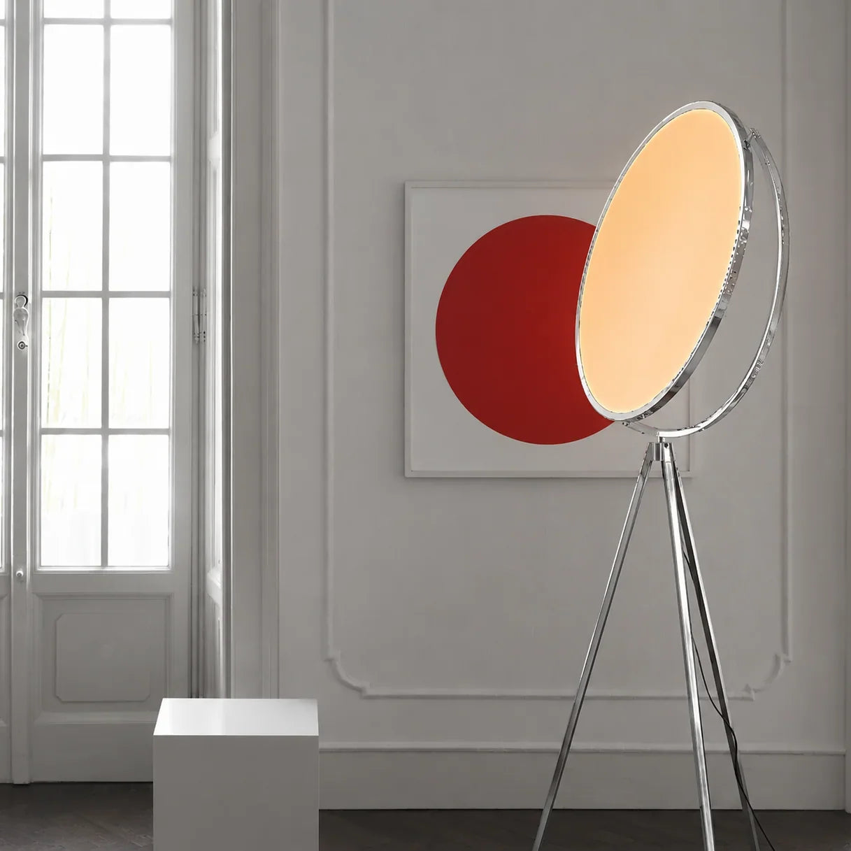 A modern tripod floor lamp with a circular shade stands next to a white wall featuring a framed artwork with a large red circle. A small white cube is positioned on the floor nearby.