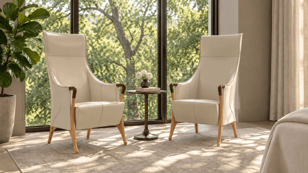 Two cream-colored armchairs with wooden legs face each other, separated by a small round table with a vase of flowers, set near large windows overlooking green trees.