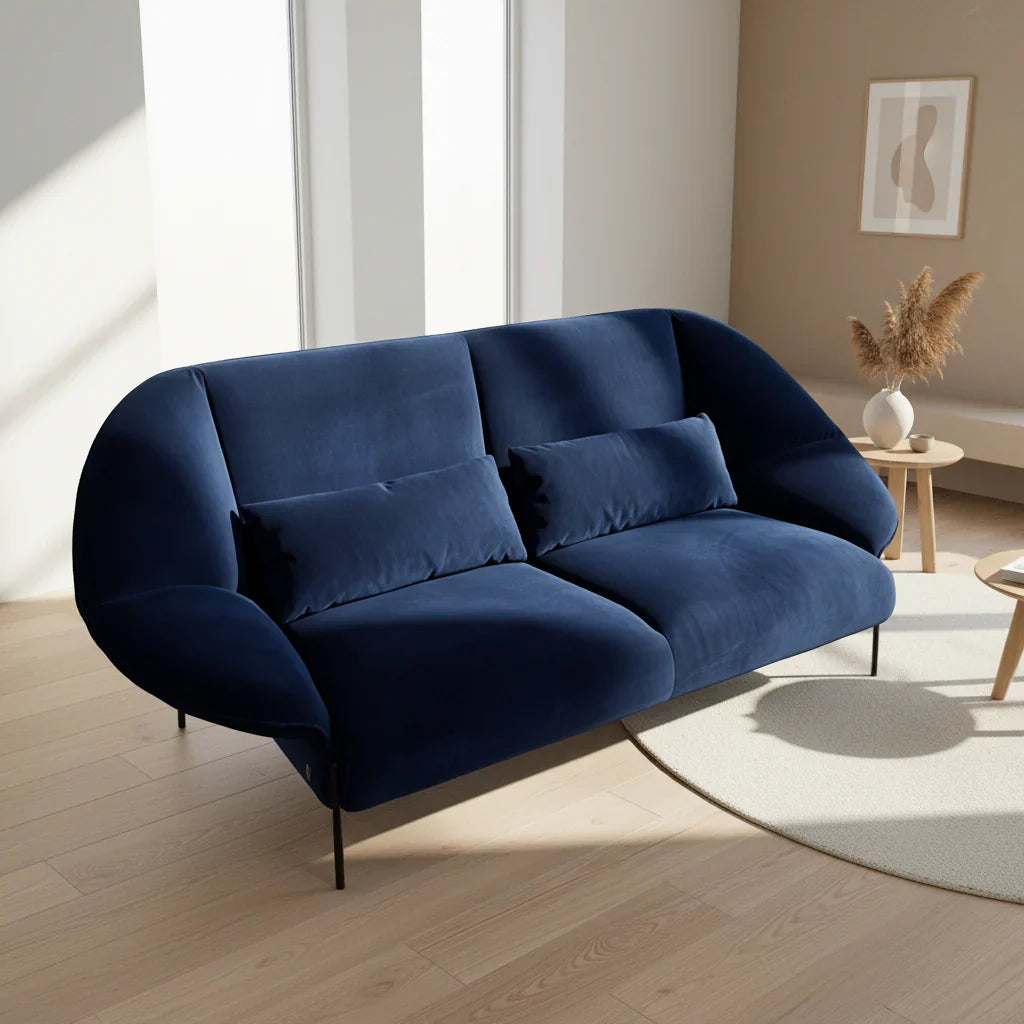 A modern blue velvet sofa with two matching cushions stands on a light wood floor in a minimalist living room with neutral decor and natural light.
