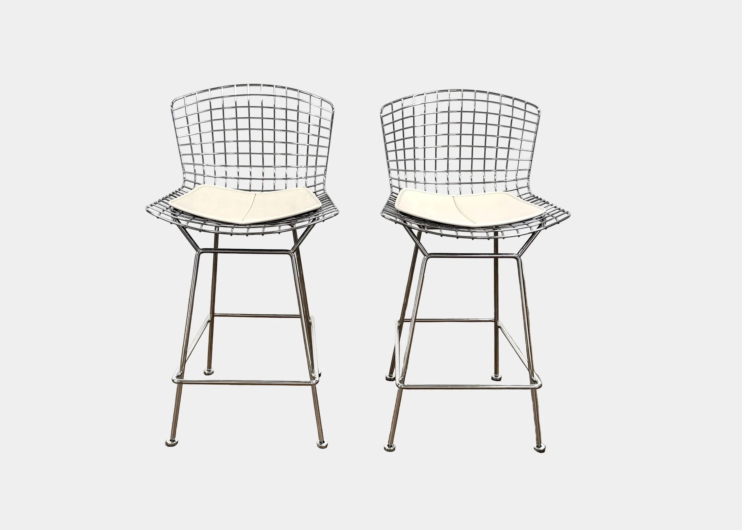 Two metal wire bar stools with white seat cushions, featuring grid-pattern backs and legs with footrests, set against a plain white background.