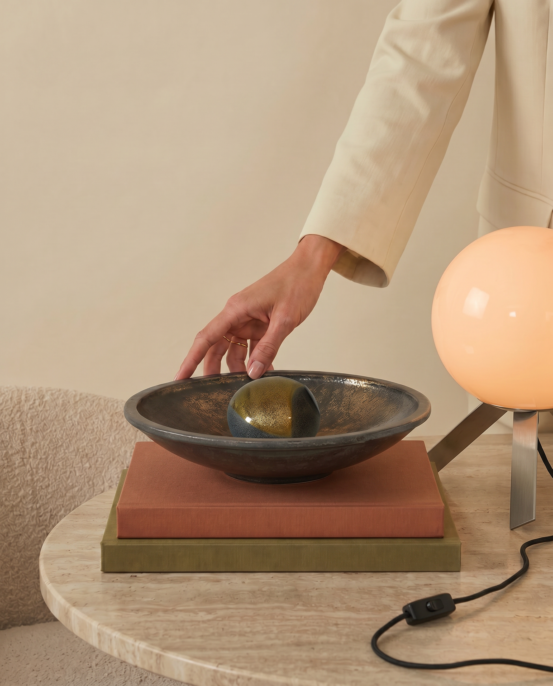 A person in a cream suit touches a metallic sphere in a shallow bowl on a stack of books, beside a round lamp on a marble table.