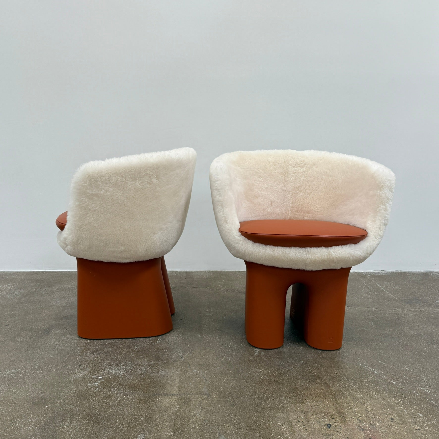 The Louis Vuitton Dolls Fur by Raw Edges is a modern chair with a curved white fluffy leather and fur seat, two short cylindrical brown legs, and a matching brown base, designed by Louis Vuitton and set against a plain background.