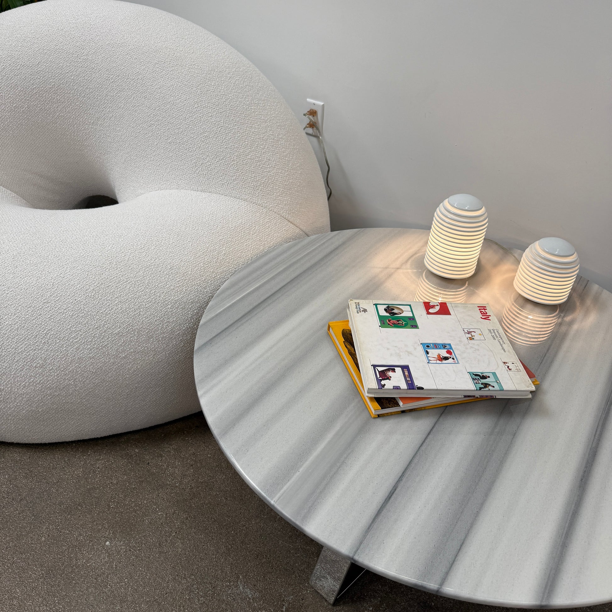 The Objects of Common Interest Tube Chair by Objects of Common Interest is a modern, light grey lounge chair with a donut-shaped design, rounded form, and central indentation, shown against a plain white background.