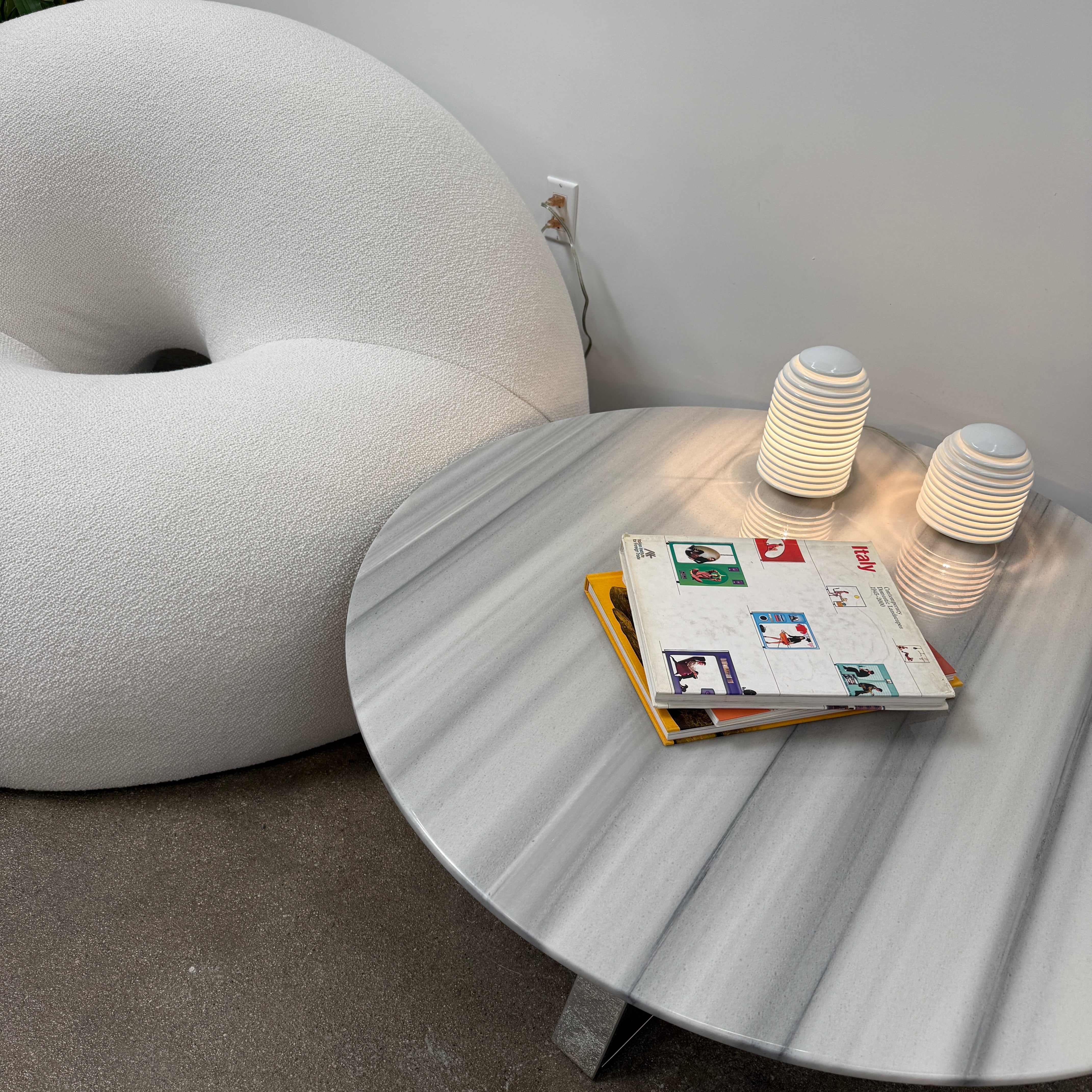 The Objects of Common Interest Tube Chair by Objects of Common Interest is a modern, light grey lounge chair with a donut-shaped design, rounded form, and central indentation, shown against a plain white background.
