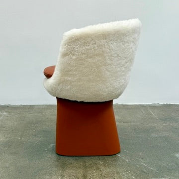 The Louis Vuitton Dolls Fur by Raw Edges is a modern chair with a curved white fluffy leather and fur seat, two short cylindrical brown legs, and a matching brown base, designed by Louis Vuitton and set against a plain background.