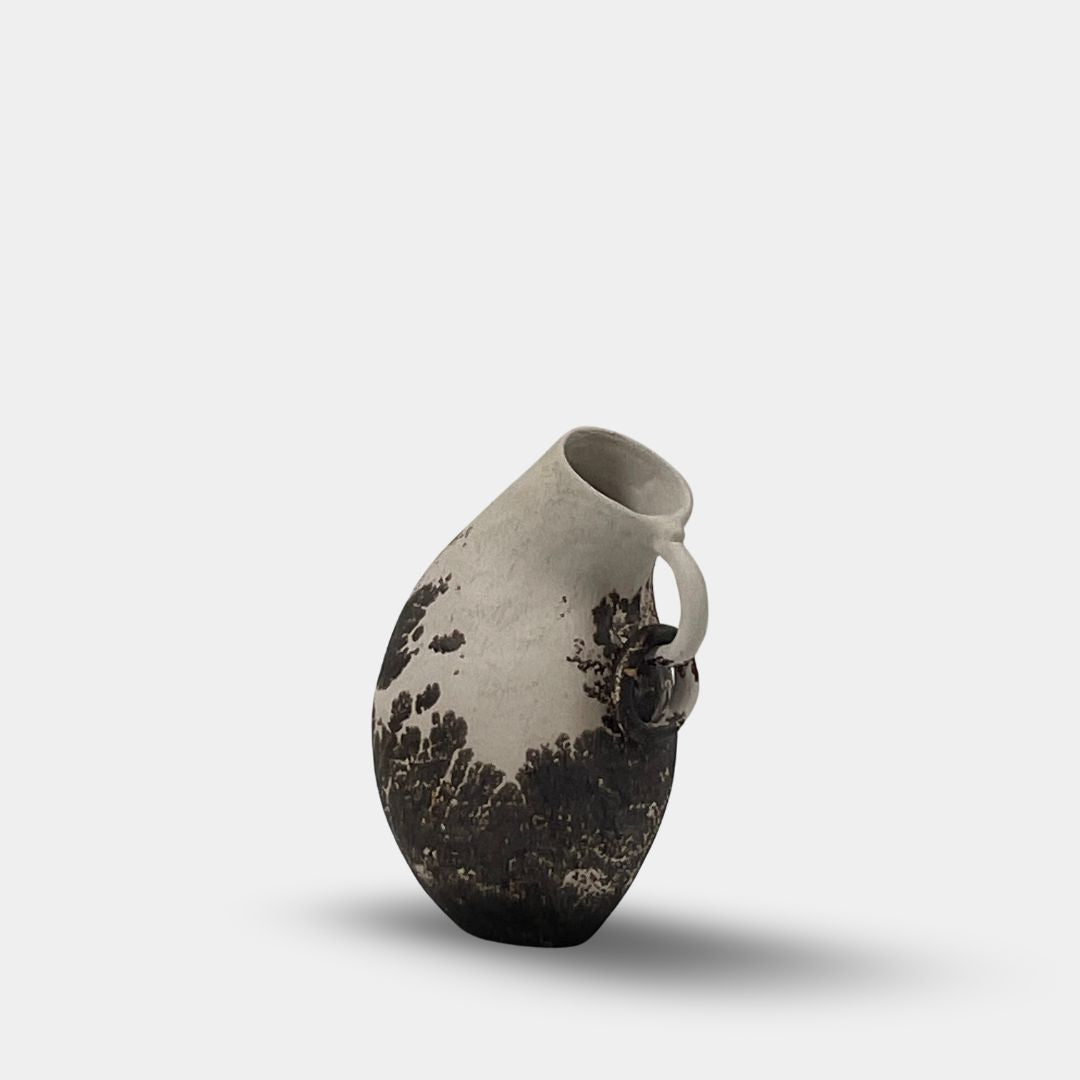 The Sofi Ceramic Curved Jug by Sofi Ceramics is a handbuilt white and black ceramic vase with an irregular shape, small handle, and abstract dark patterns reminiscent of sculptured vessels.