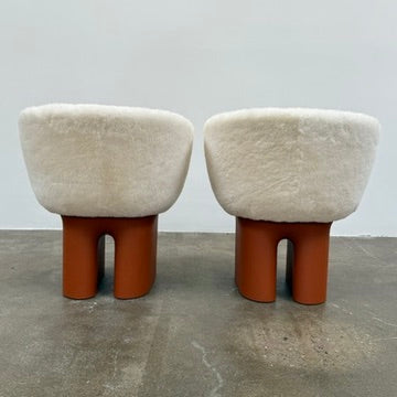 The Louis Vuitton Dolls Fur by Raw Edges is a modern chair with a curved white fluffy leather and fur seat, two short cylindrical brown legs, and a matching brown base, designed by Louis Vuitton and set against a plain background.