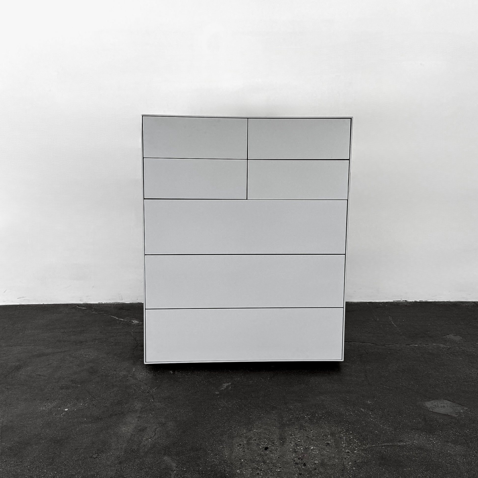 The Treku Lauki Tall Dresser by Treku, with a sleek lacquered finish and push-latch drawers—one partially open—stands on a concrete floor against a plain white wall.