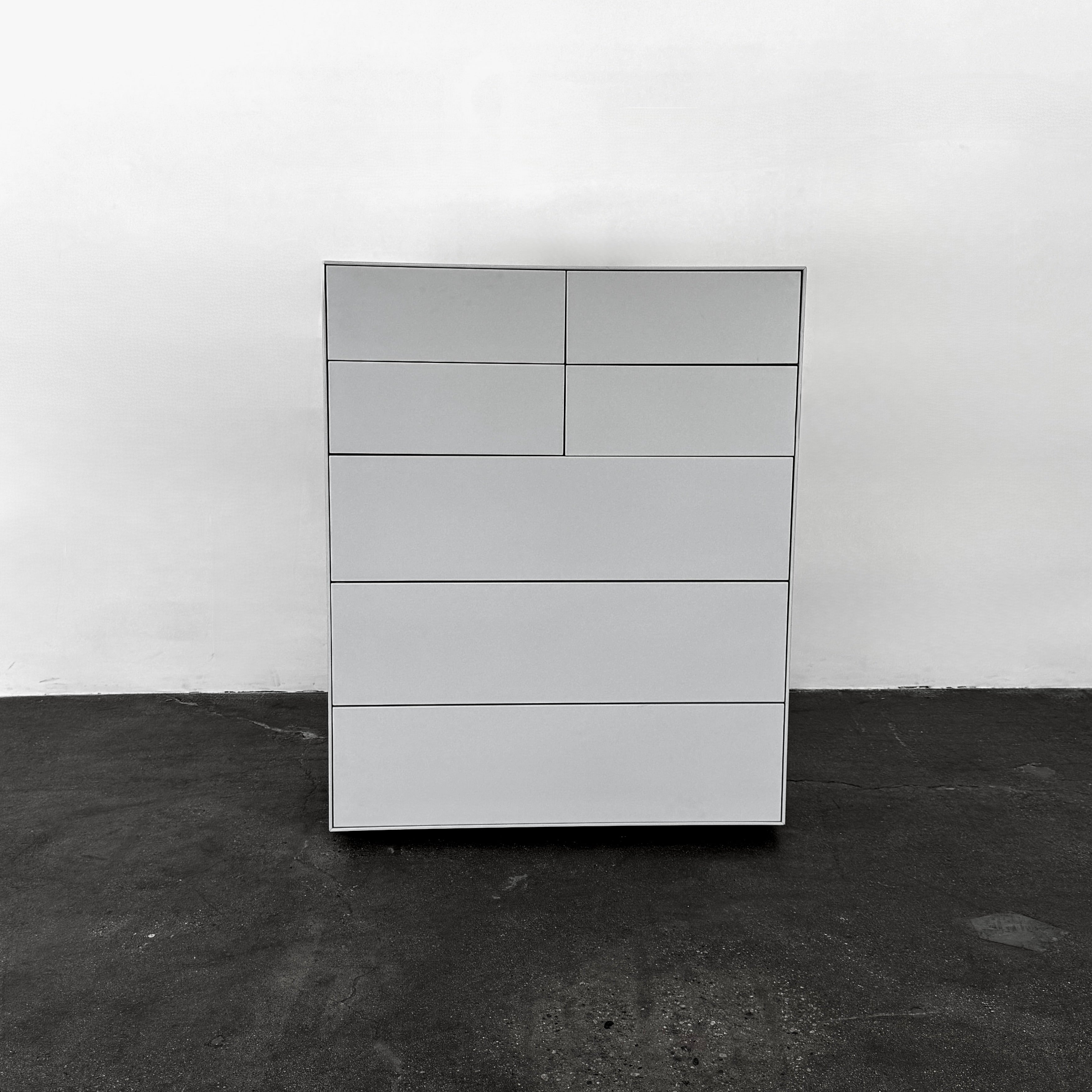 The Treku Lauki Tall Dresser by Treku, with a sleek lacquered finish and push-latch drawers—one partially open—stands on a concrete floor against a plain white wall.