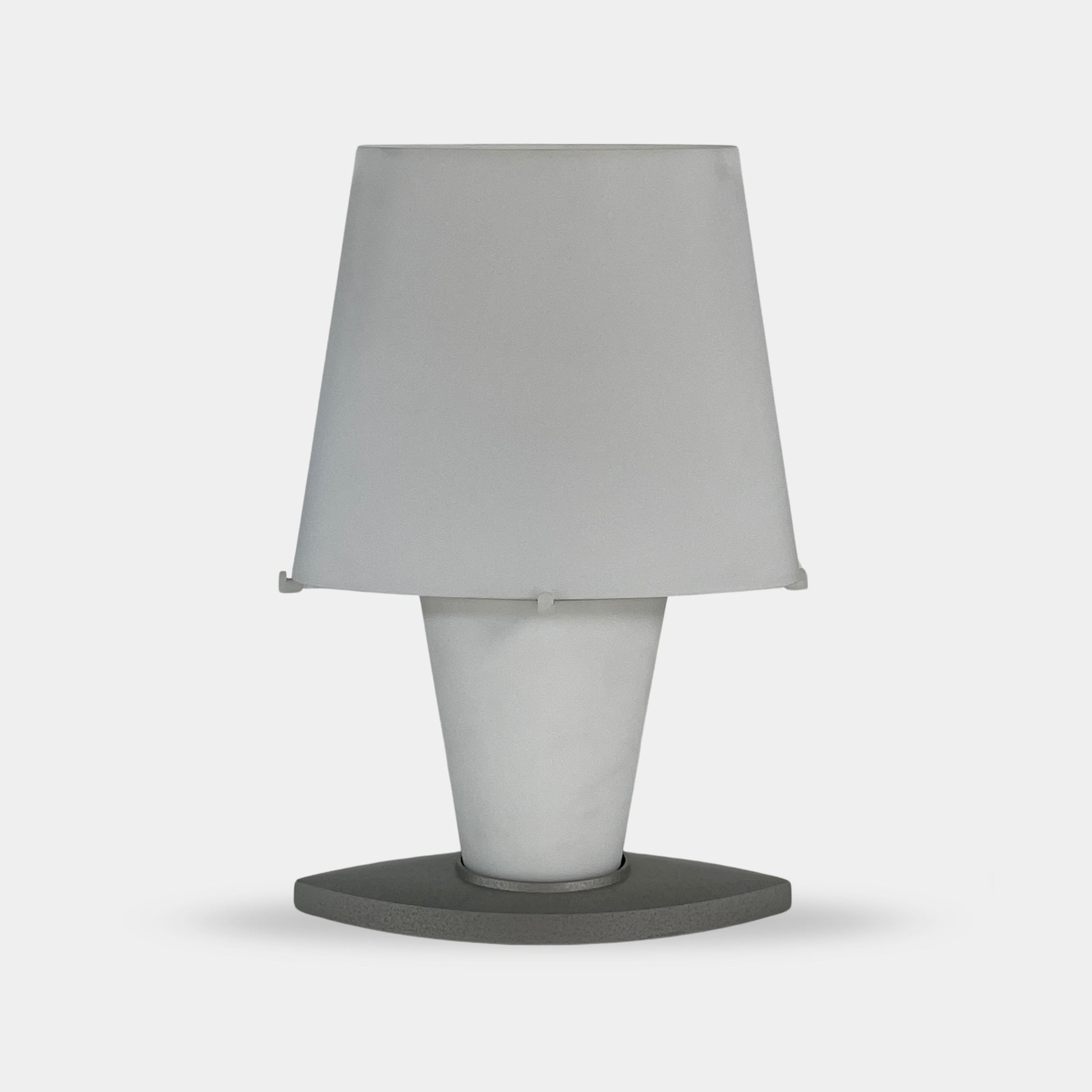 The Fontana Arte 3064 Table Light by Fontana Arte, echoing Daniela Puppa's designs, boasts a wide conical shade for a warm glow. It sits elegantly on a white surface against a plain backdrop, capturing the essence of postmodern art.