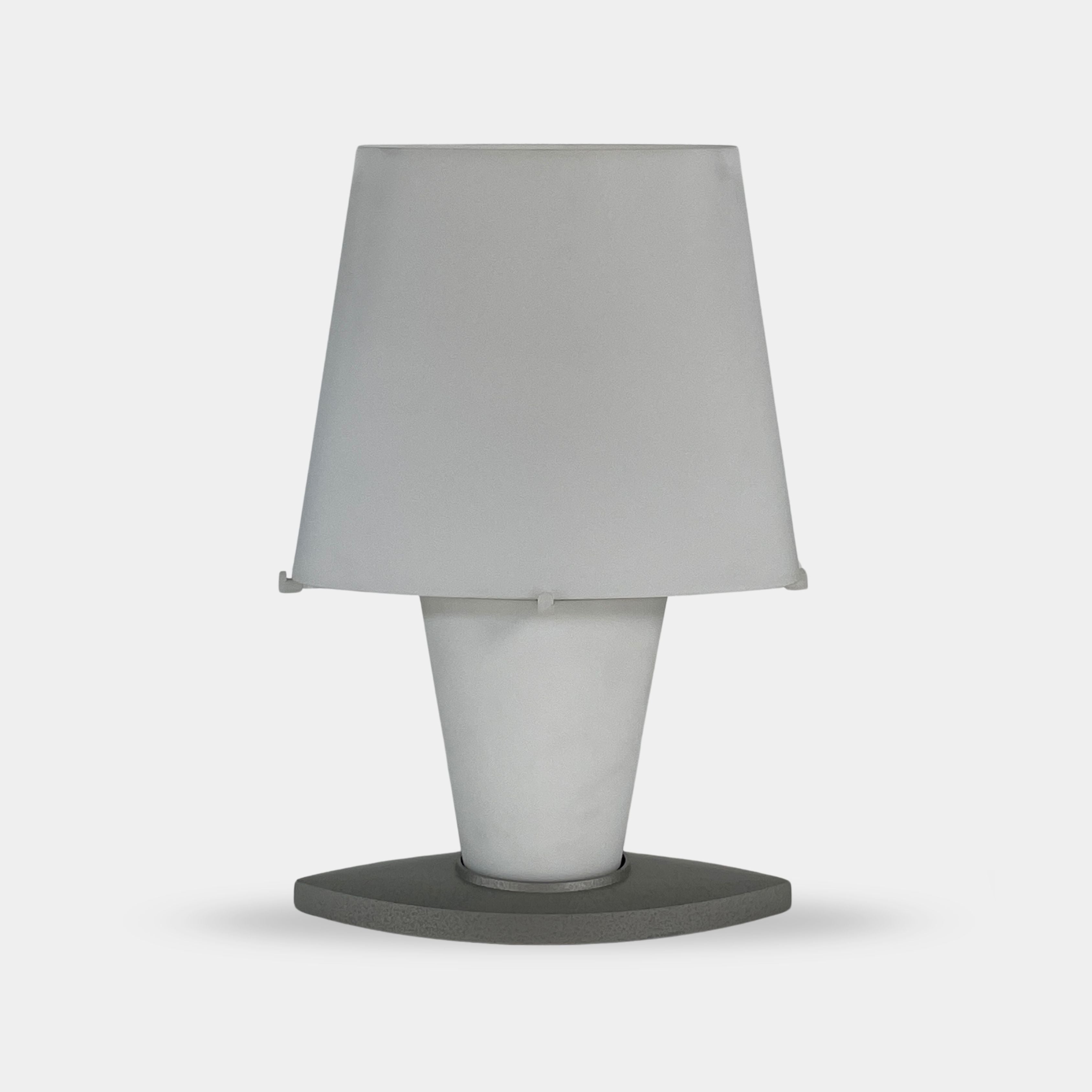 The Fontana Arte 3064 Table Light by Fontana Arte, echoing Daniela Puppa's designs, boasts a wide conical shade for a warm glow. It sits elegantly on a white surface against a plain backdrop, capturing the essence of postmodern art.