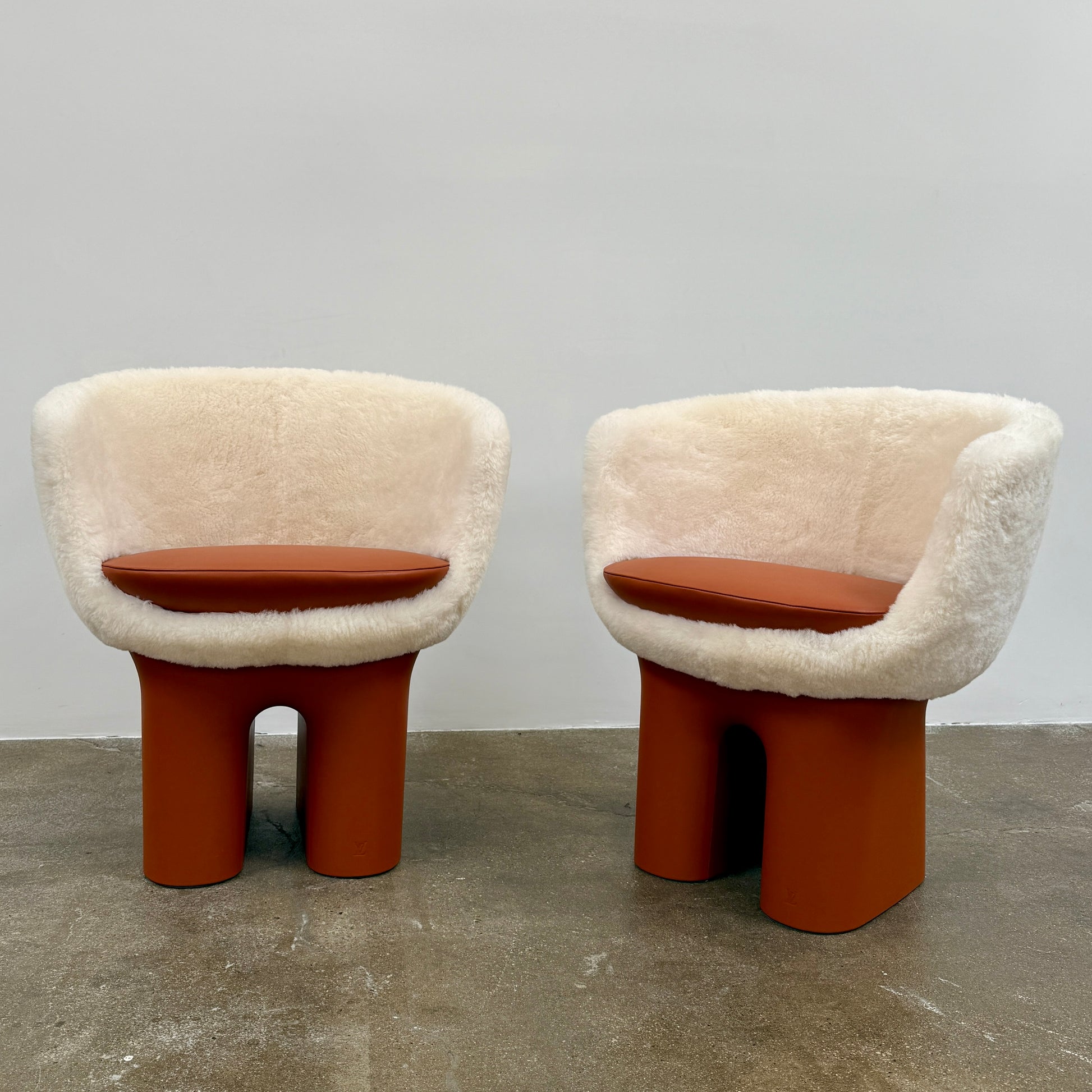 The Louis Vuitton Dolls Fur by Raw Edges is a modern chair with a curved white fluffy leather and fur seat, two short cylindrical brown legs, and a matching brown base, designed by Louis Vuitton and set against a plain background.