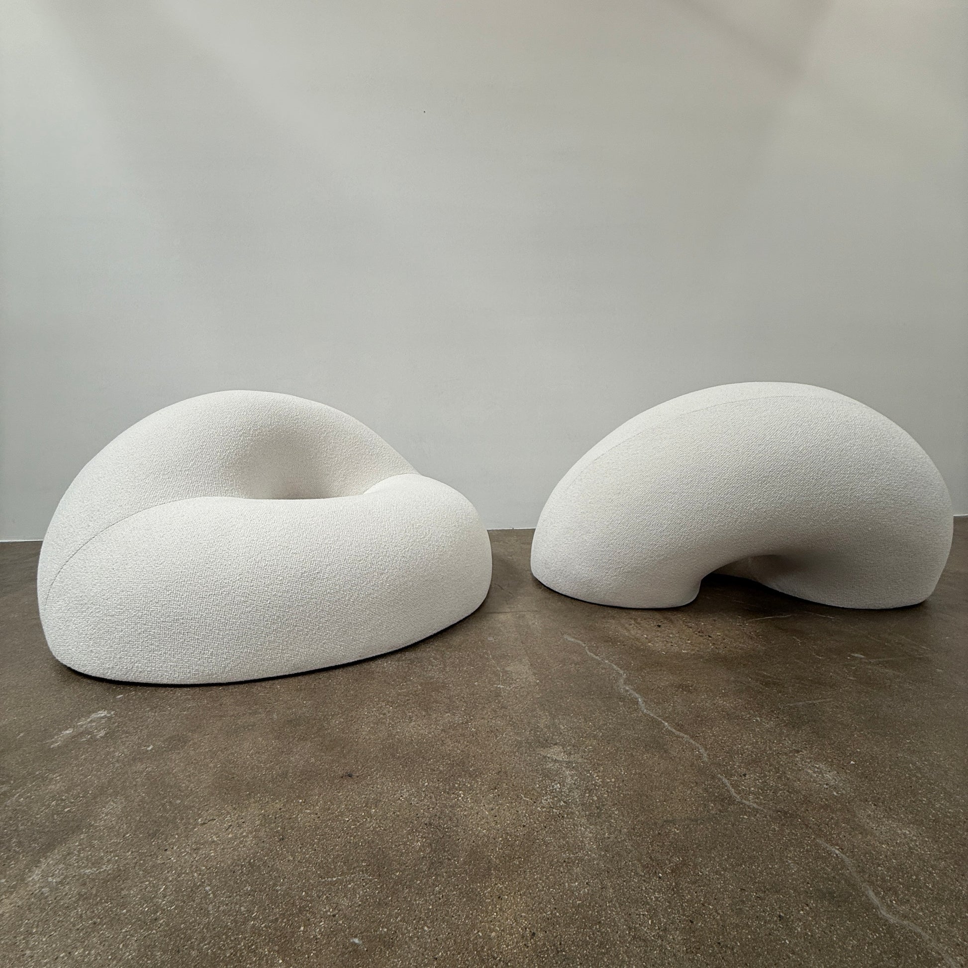The Objects of Common Interest Tube Chair by Objects of Common Interest is a modern, light grey lounge chair with a donut-shaped design, rounded form, and central indentation, shown against a plain white background.