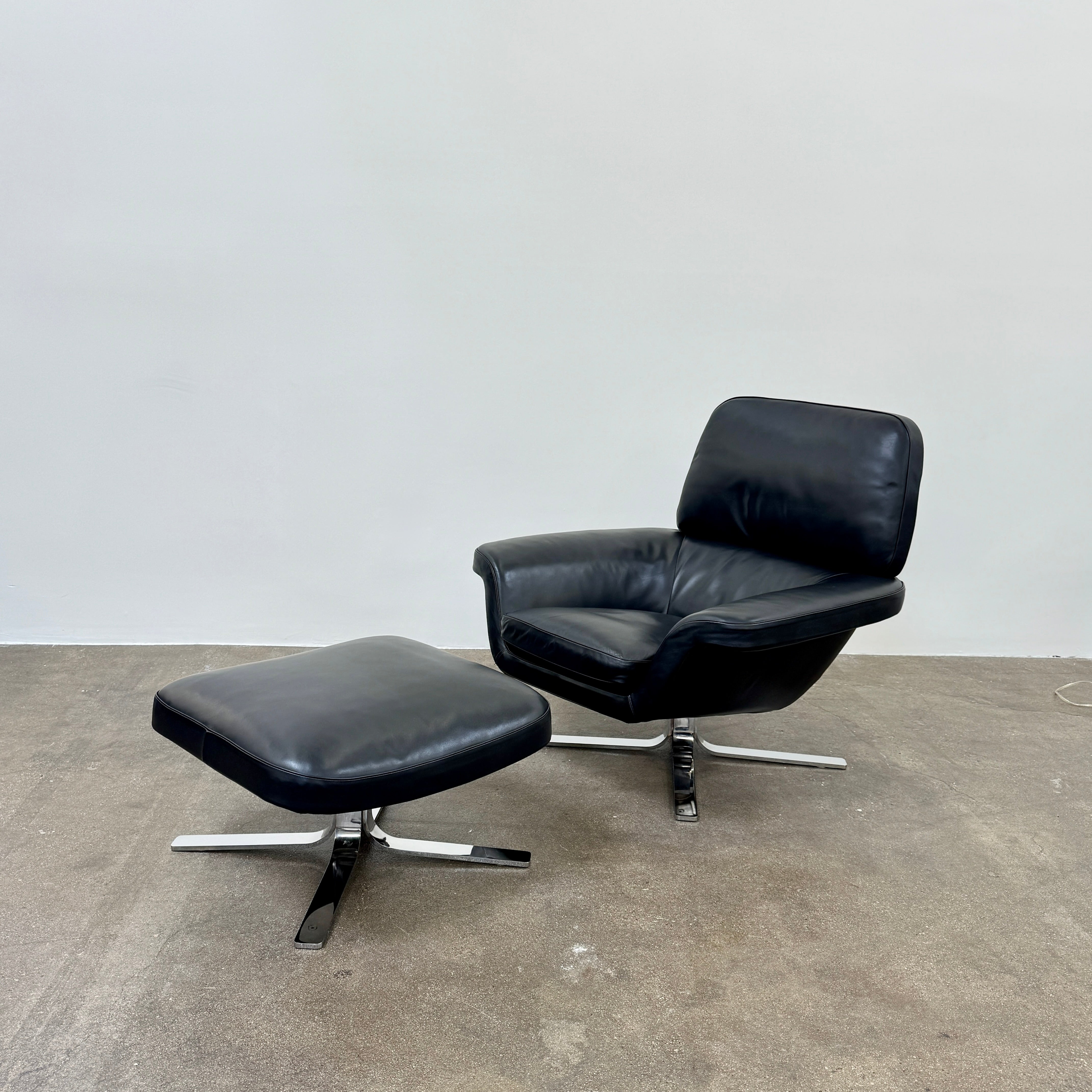 A Minotti Blake Lounge Chair & Ottoman by Minotti in black leather with chrome swivel bases, positioned on a bare concrete floor against a plain light-colored wall.