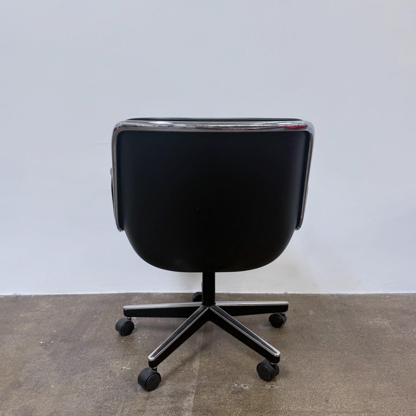 A DWR Pollock Executive Chair with a chrome frame and five wheels is shown from the back, set on a concrete floor against a plain white wall.