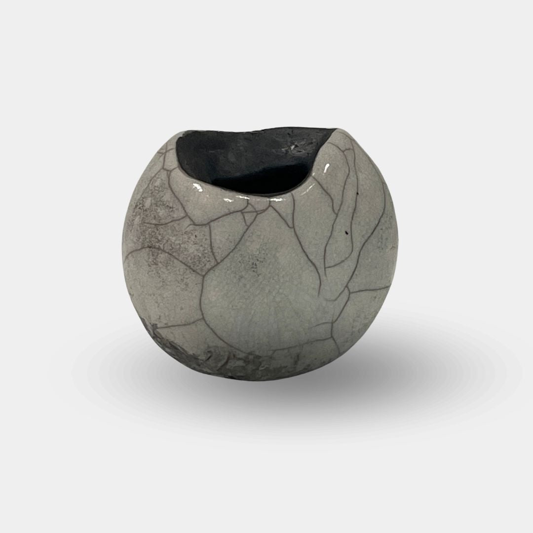 The Sofi Ceramic Raku Small Vessel by Sofi Ceramics is a round, grey ceramic vase with a marble crack-like pattern, drawing inspiration from textured African cultural vessels, and it features a wide, uneven opening against a plain white background.