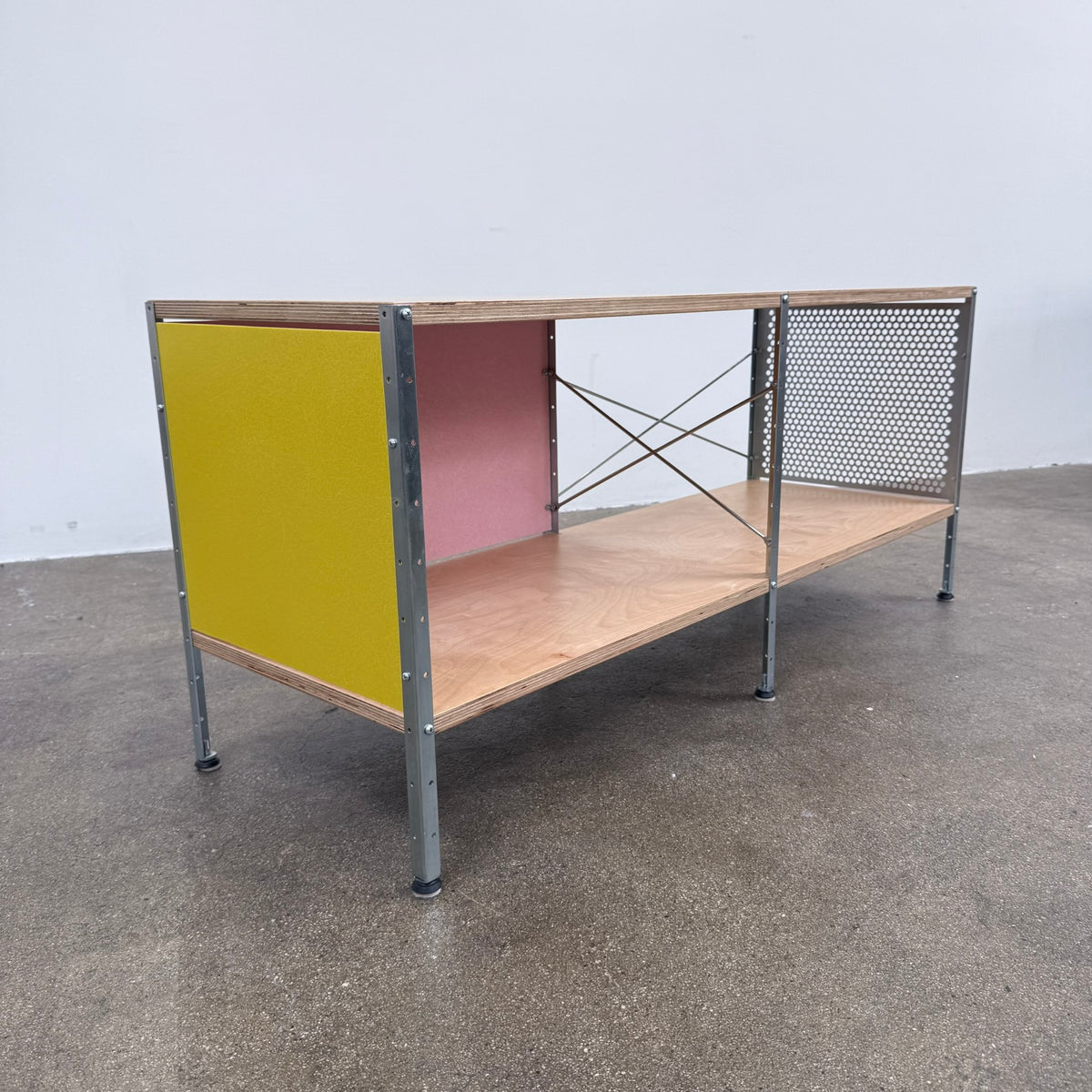 Herman Miller Eames Storage Unit featuring a low rectangular frame with plywood shelves, a yellow side panel, a pink back panel, and a perforated metal side.