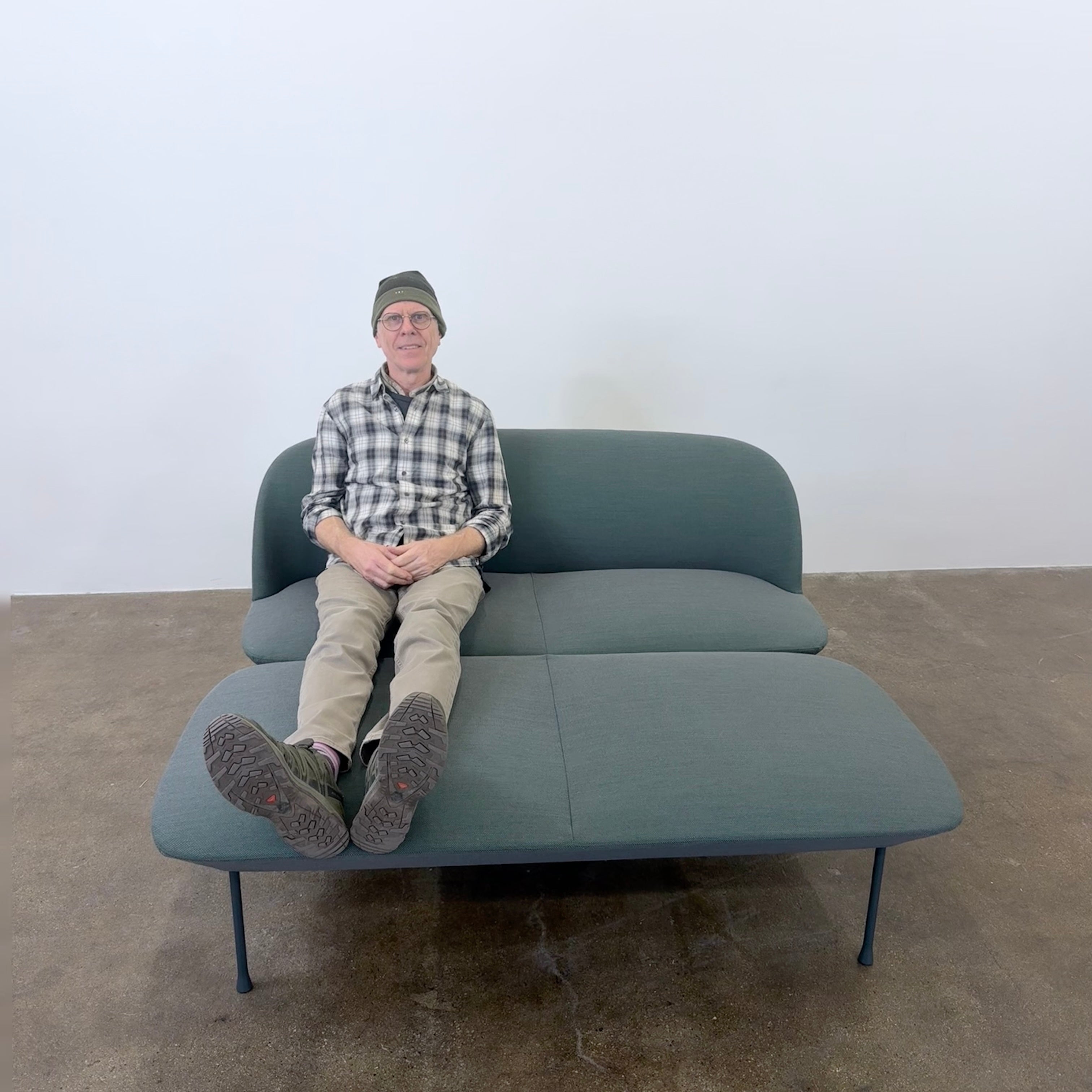 The Muuto Oslo 2-seat sofa & bench by Muuto, featuring modern green upholstery and slim black legs inspired by Danish design, stands in a bright room with beige walls and sheer white curtains.