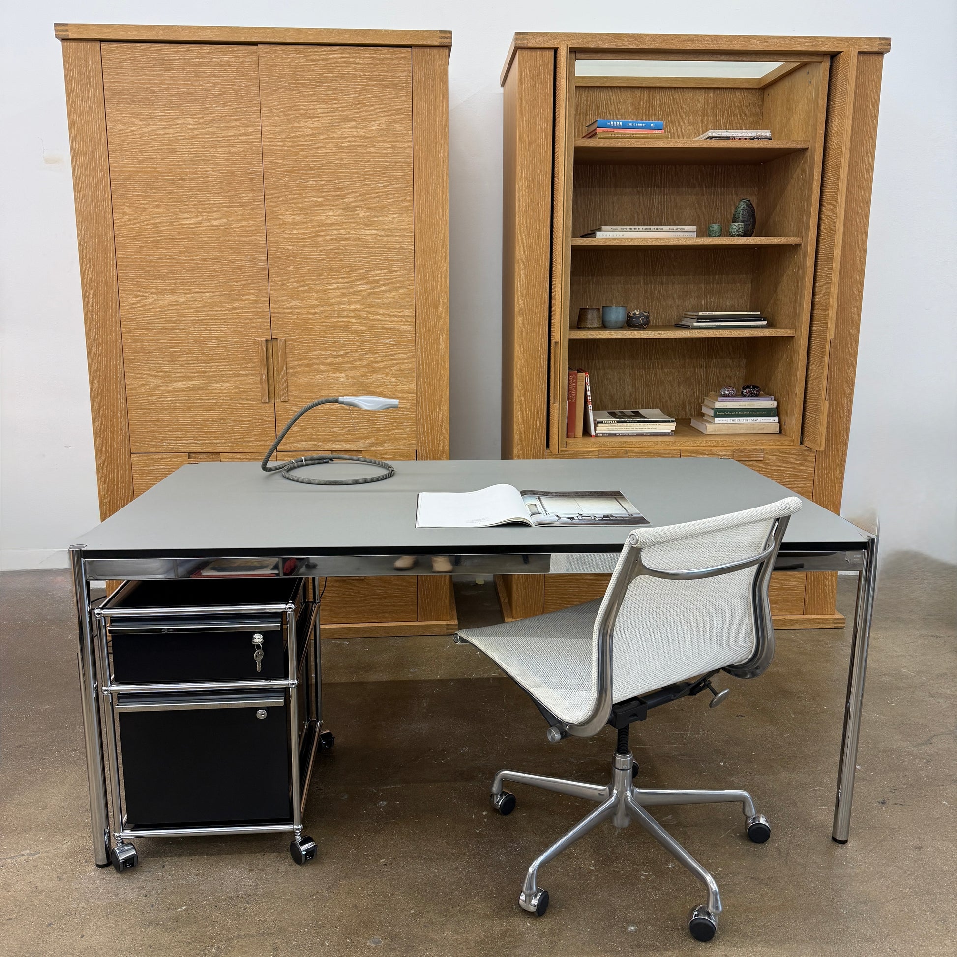 A modern mesh-back office chair sits at a USM Haller Work Table by USM Haller, which has a minimalist design and metal legs, placed on a concrete floor with a white background.