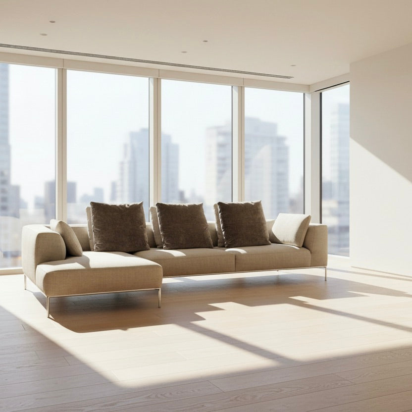 The B&B Italia Frank Sectional is a modern beige sofa featuring three large gray cushions, one beige side cushion, and sleek metal legs—an elegant statement piece from B&B Italia, set off by a plain white wall and concrete floor.