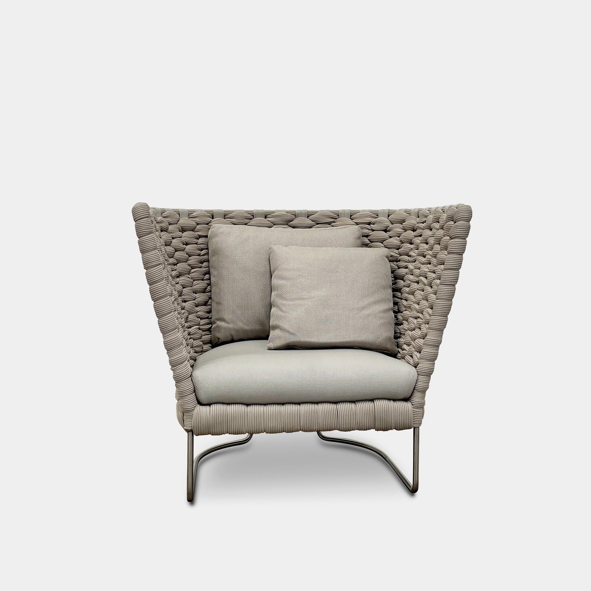 The Paola Lenti Ami Outdoor Armchair by Paola Lenti features a large weave frame, light gray cushions, and a metal base, all set against a plain white background.