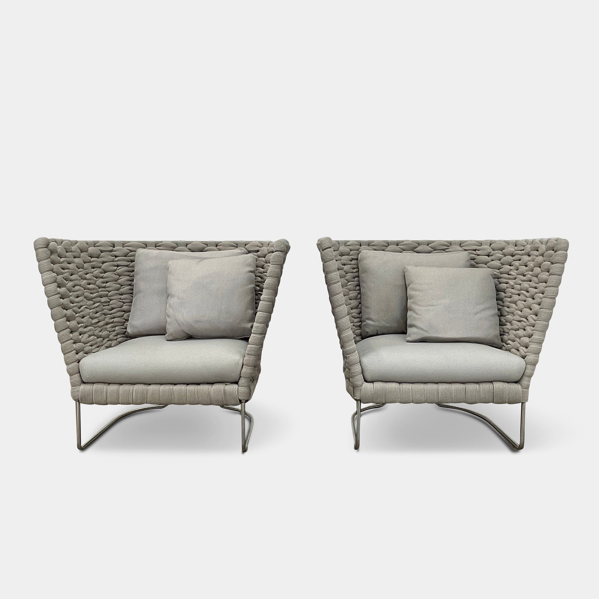 The Paola Lenti Ami Outdoor Armchair by Paola Lenti features a large weave frame, light gray cushions, and a metal base, all set against a plain white background.