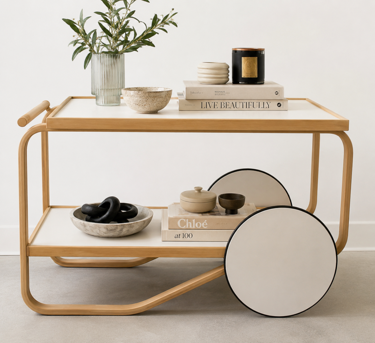 The Artek Aalto Tea Trolley 901 by Artek is a wooden bar cart inspired by the iconic Aalto Tea Trolley, showcasing two large white wheels, a handle, and two white shelves. Set against a plain white background, it stands as a testament to handcrafted Finnish design in the spirit of Alvar Aalto.