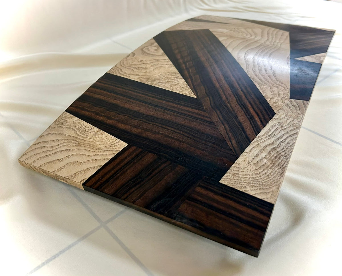 A rectangular table top storage box by Mike Fair Custom Furnishings, crafted from luxurious Macassar Ebony and Ash Burl with a geometric patterned lid, is displayed open to reveal a foam insert inside. The elegant Mike Fair Table Top Storage Box rests on a light fabric surface.