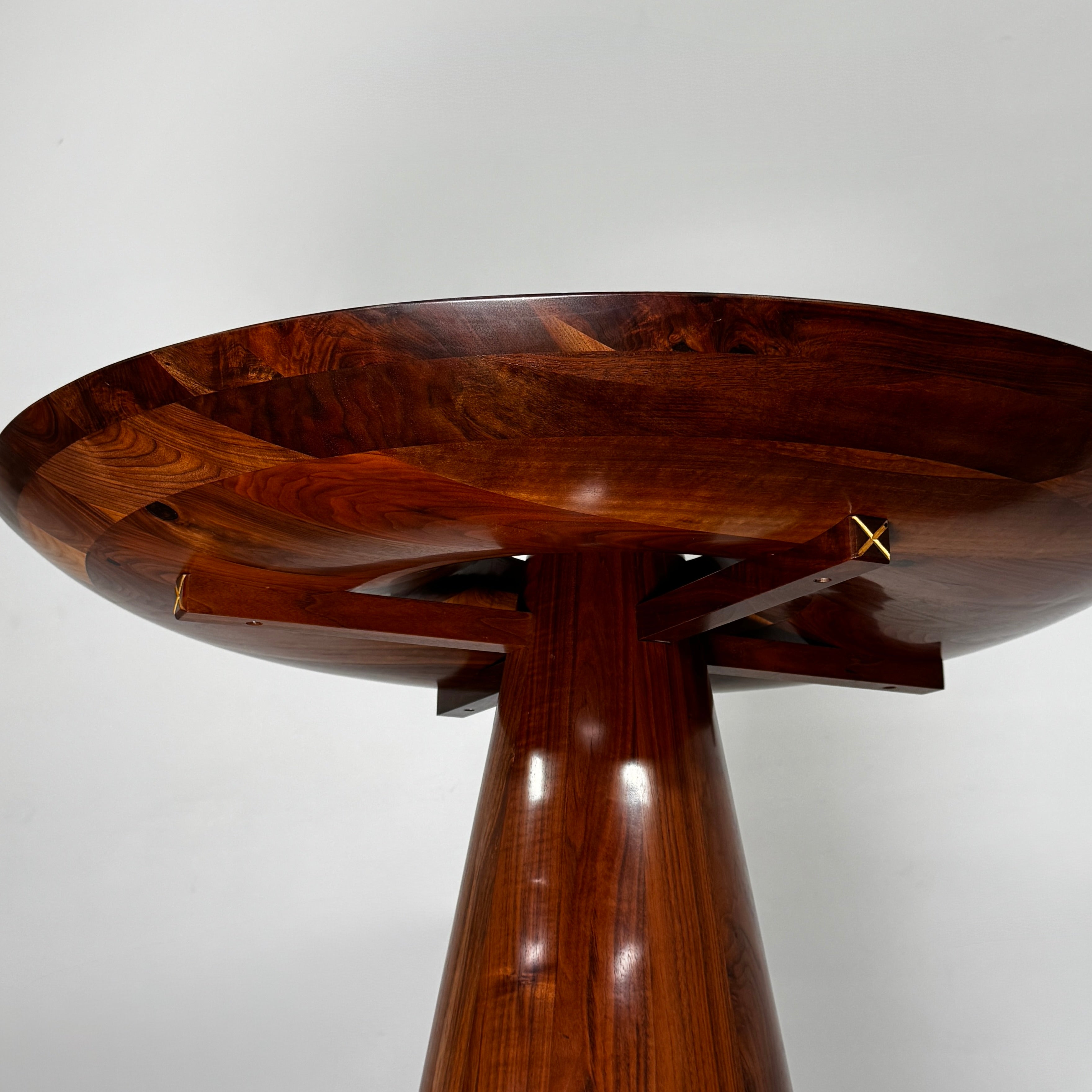 The Mike Fair Claro Walnut Bar Height Table by Artist features a round claro walnut top with a central circular cutout, a conical base, and a small gold cross detail on the side, shown on a concrete floor.
