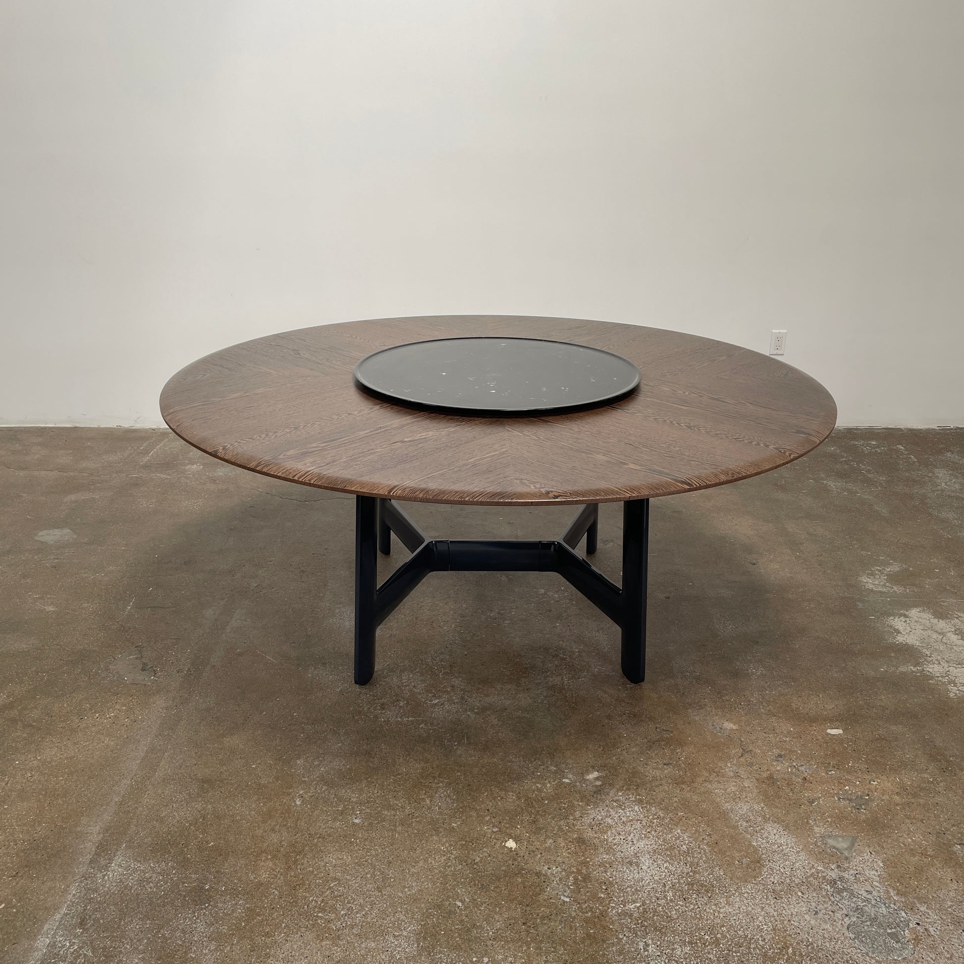 Round wooden table with a central raised circular platform and sleek black legs, reminiscent of the elegant design seen in the B&B Italia Alex Dining Table With Rotating Marble Tray. The rotating marble tray adds a refined touch, making it a versatile centerpiece on a white background.