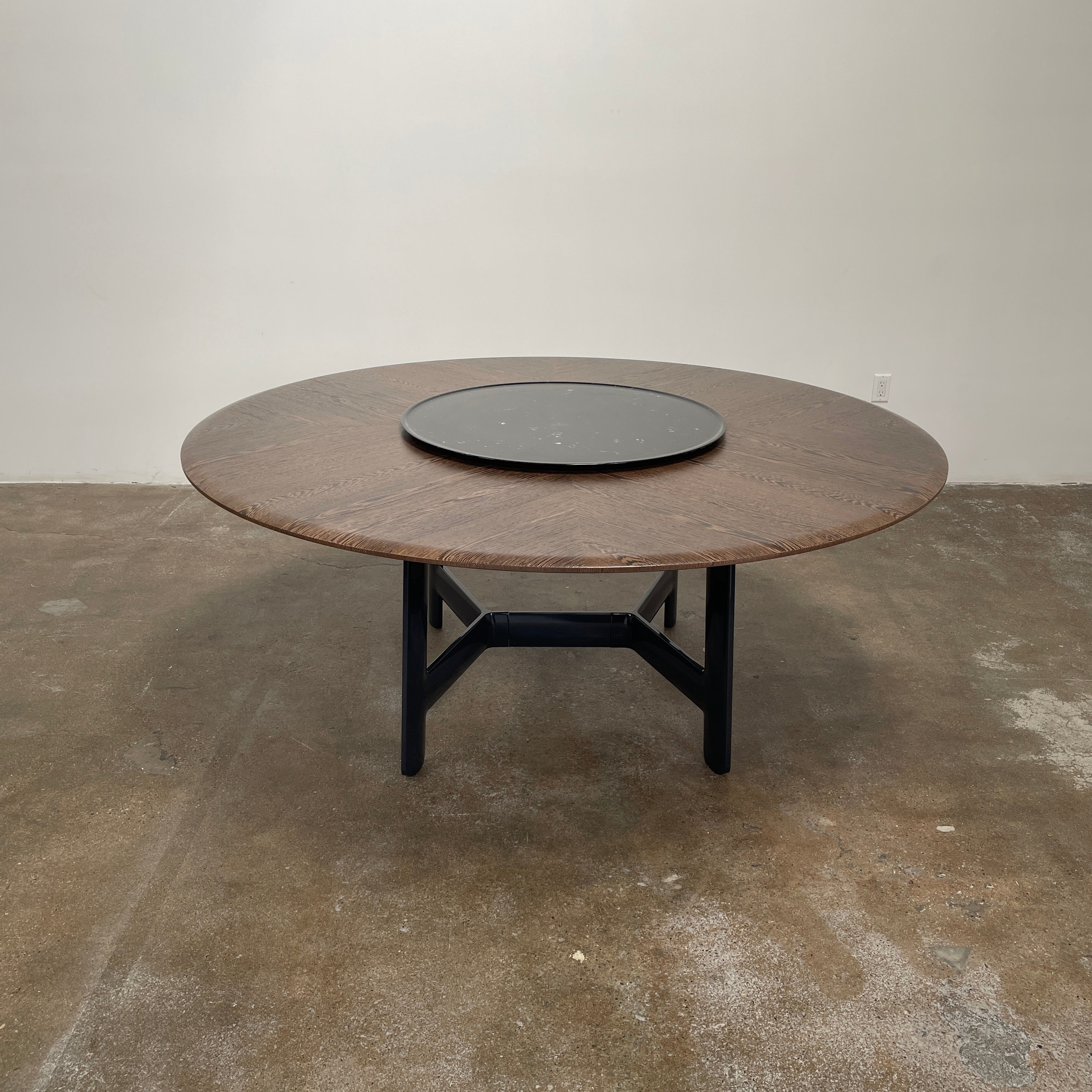 Round wooden table with a central raised circular platform and sleek black legs, reminiscent of the elegant design seen in the B&B Italia Alex Dining Table With Rotating Marble Tray. The rotating marble tray adds a refined touch, making it a versatile centerpiece on a white background.