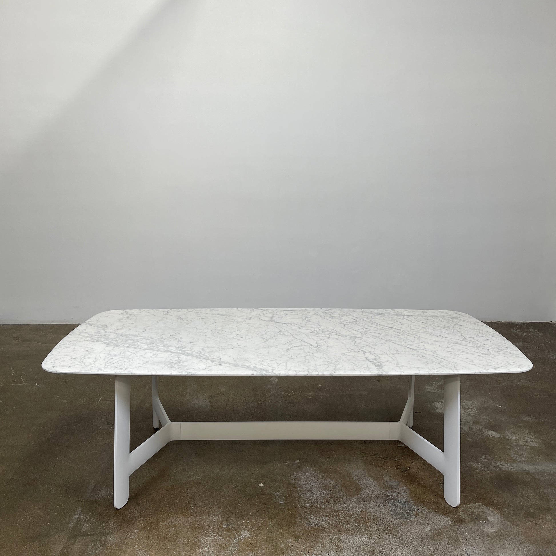 On a concrete floor against a plain white wall stands the B&B Italia Matte Carrara Marble Alex Dining Table, showcasing contemporary design with its long, rectangular form and white base, adding both style and sophistication to the space.