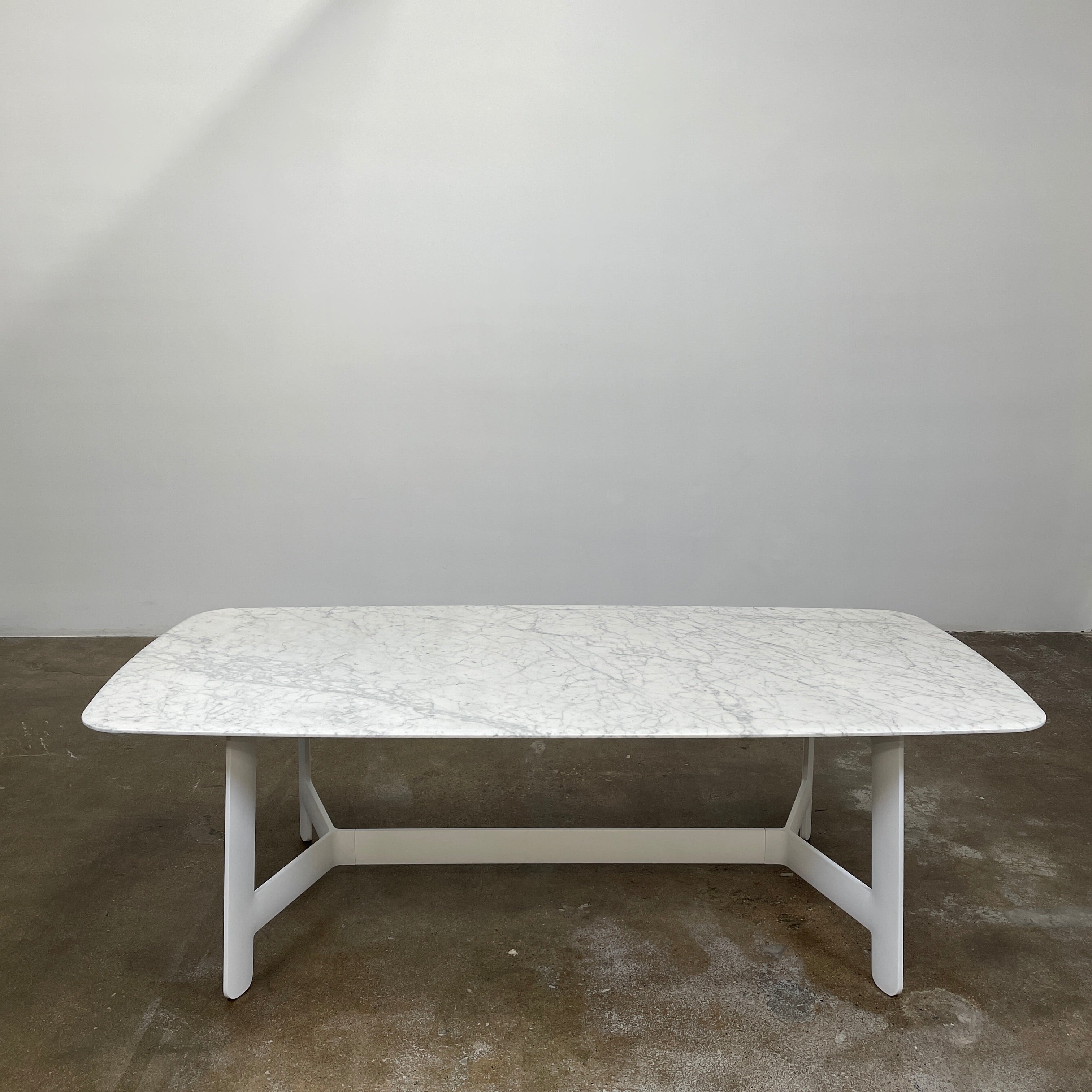 On a concrete floor against a plain white wall stands the B&B Italia Matte Carrara Marble Alex Dining Table, showcasing contemporary design with its long, rectangular form and white base, adding both style and sophistication to the space.