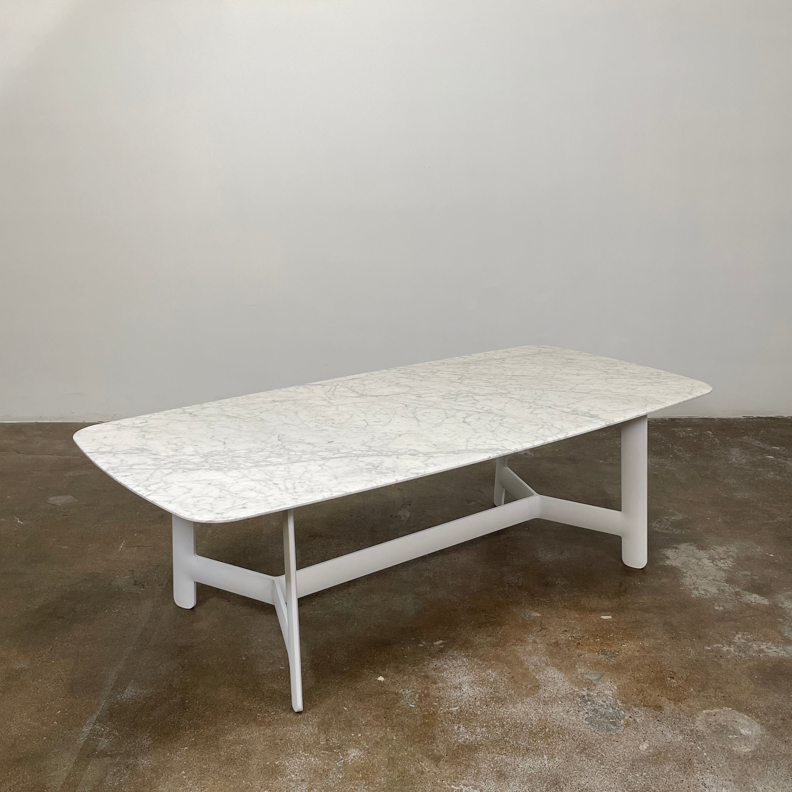 On a concrete floor against a plain white wall stands the B&B Italia Matte Carrara Marble Alex Dining Table, showcasing contemporary design with its long, rectangular form and white base, adding both style and sophistication to the space.