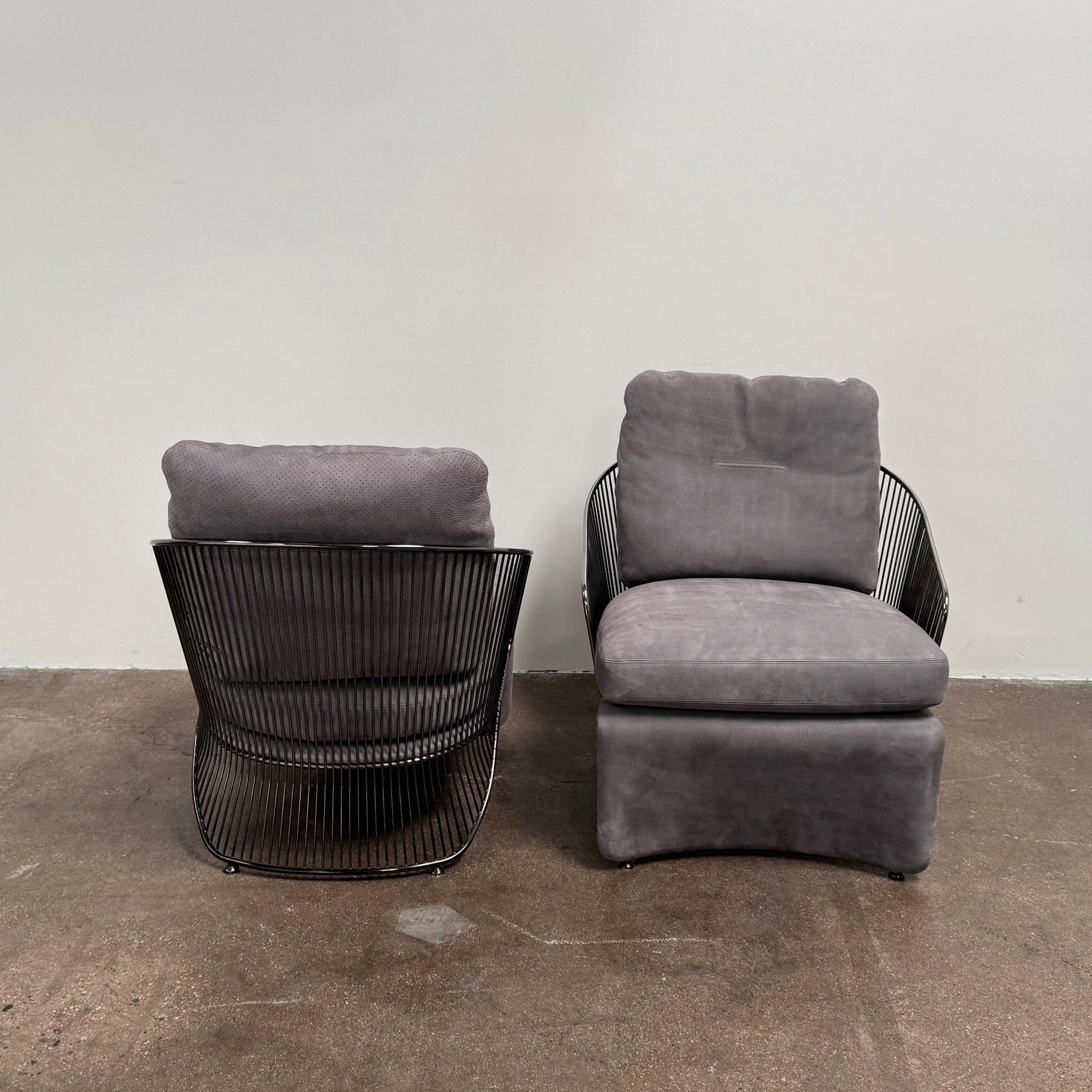 A set of two Minotti Colette Lounge Chairs by Minotti is displayed side by side against a plain white background. Each chair showcases luxurious gray nubuck leather upholstery and sleek black metal armrests, elegantly capturing the timeless mid-century silhouette characteristic of the iconic design.