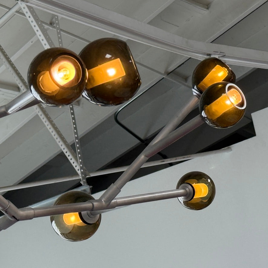 The Pouenat Last Night Branche Chandelier by Pouenat, featuring mouth-blown glass spheres, hangs beneath a white exposed beam ceiling with visible ductwork—an ideal statement for modern industrial interiors.
