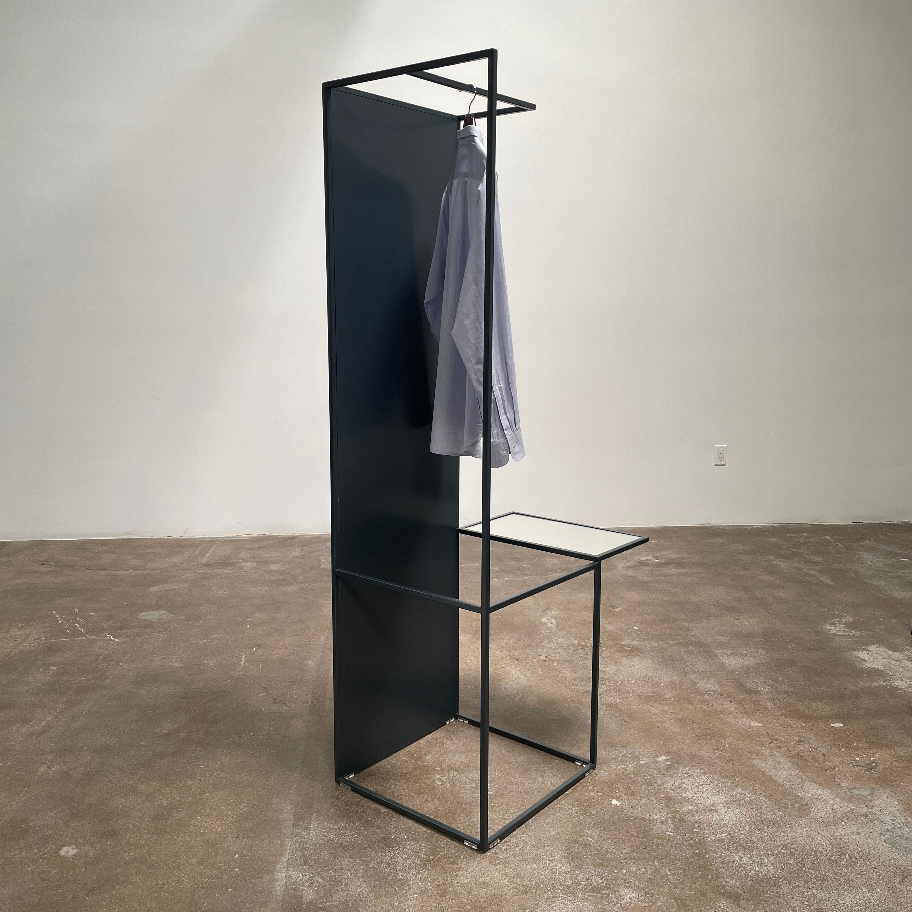 Tall, rectangular Cappellini Waku Mirrored Clothing Rack with a black metal frame, reflecting a room with a gray sofa, a colorful painting, and a beige carpet.