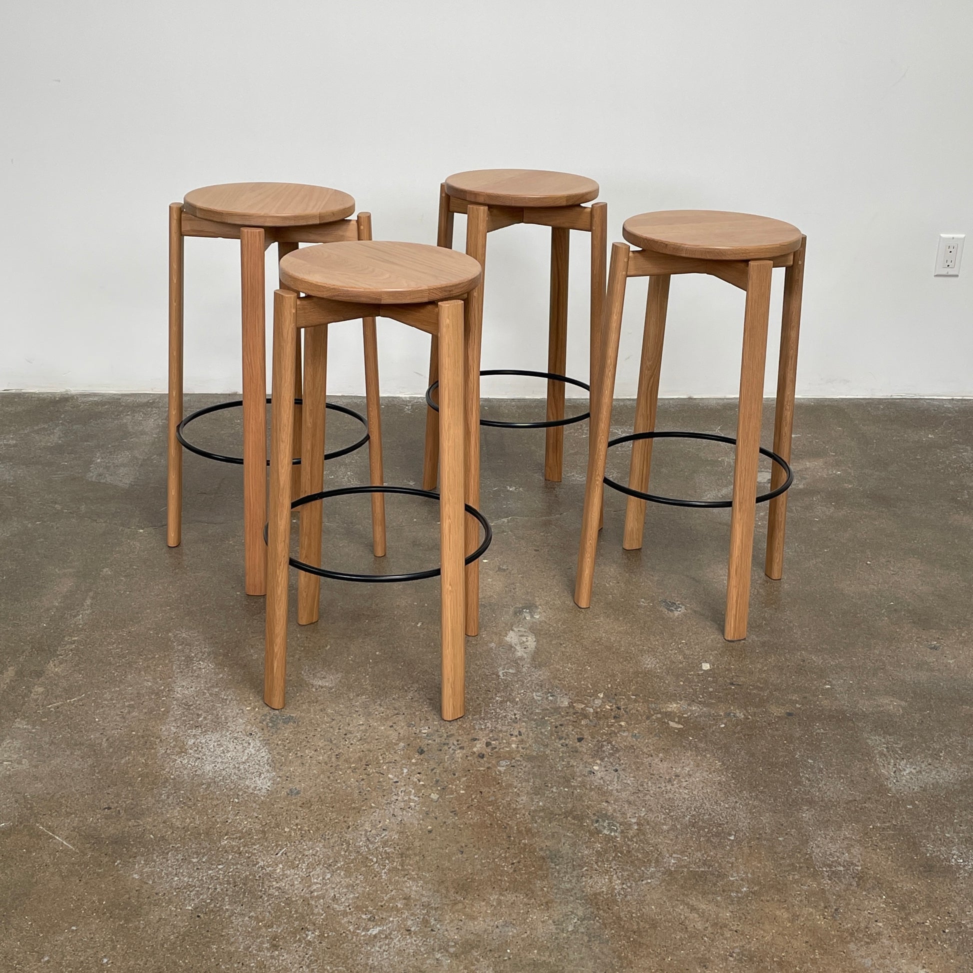 Audo Passage Bar Stools by Audo—two solid oak stools with round seats, three legs, and a black circular metal footrest—highlight Danish craftsmanship, shown against a plain white background.