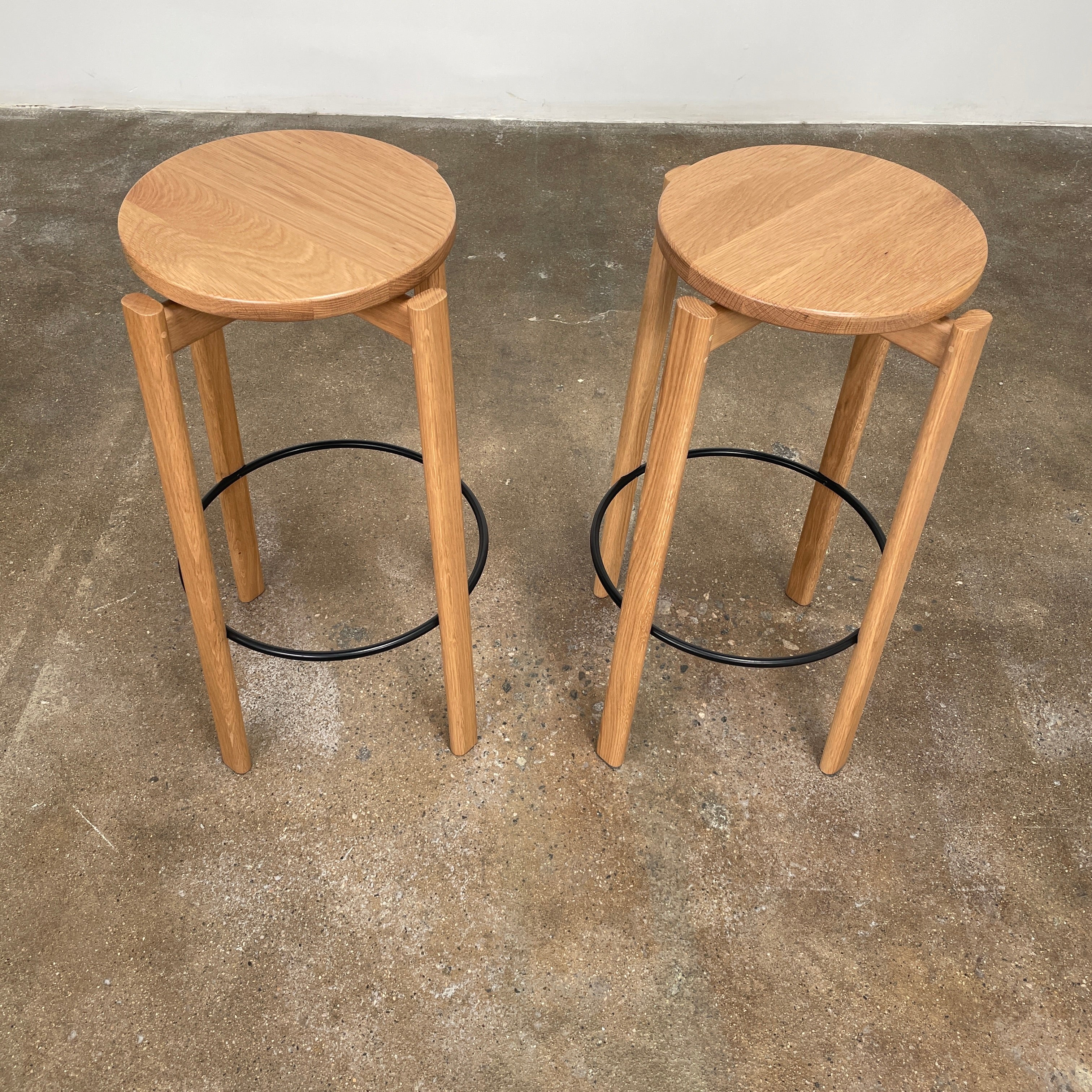 Audo Passage Bar Stools by Audo—two solid oak stools with round seats, three legs, and a black circular metal footrest—highlight Danish craftsmanship, shown against a plain white background.