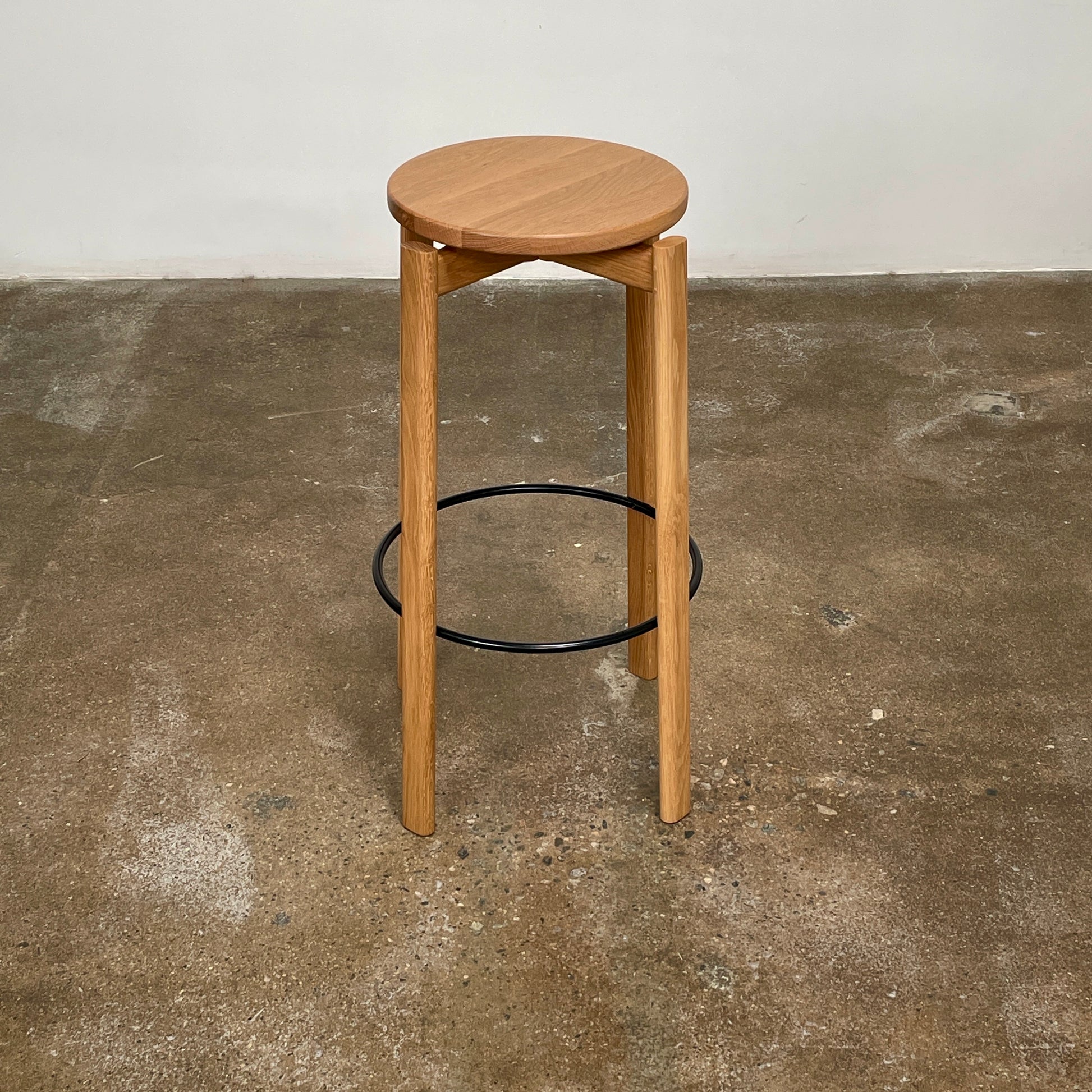 Audo Passage Bar Stools by Audo—two solid oak stools with round seats, three legs, and a black circular metal footrest—highlight Danish craftsmanship, shown against a plain white background.
