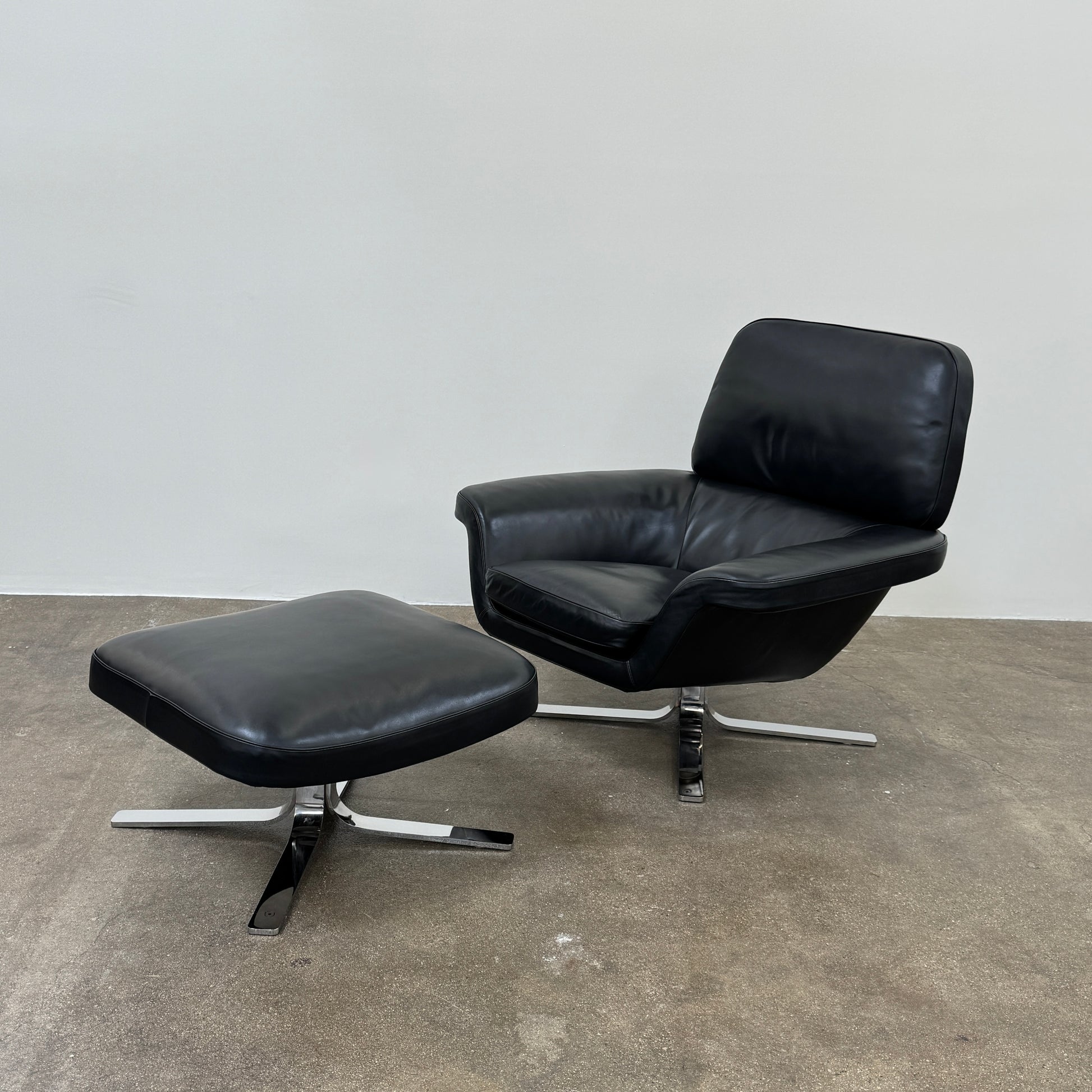 A Minotti Blake Lounge Chair & Ottoman by Minotti in black leather with chrome swivel bases, positioned on a bare concrete floor against a plain light-colored wall.