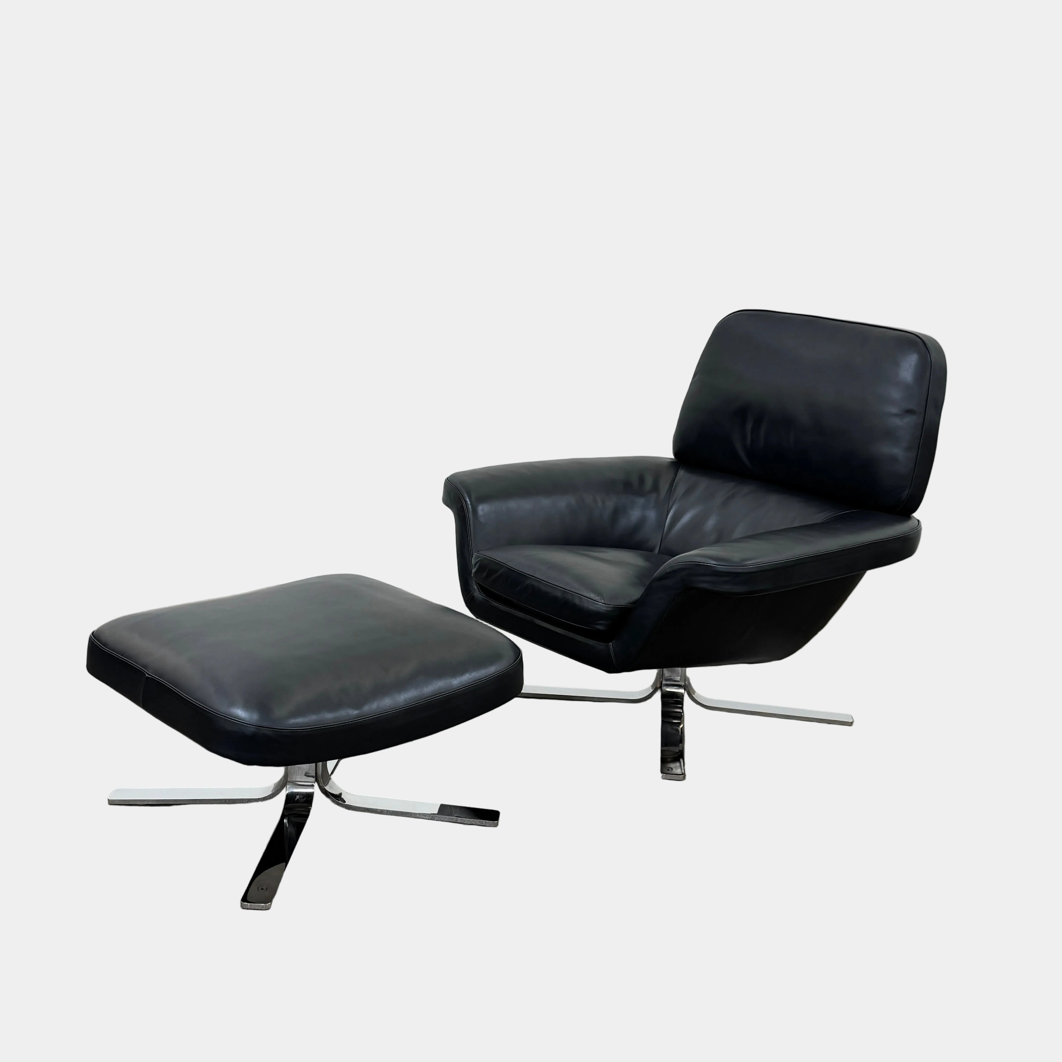 A Minotti Blake Lounge Chair & Ottoman by Minotti in black leather with chrome swivel bases, positioned on a bare concrete floor against a plain light-colored wall.