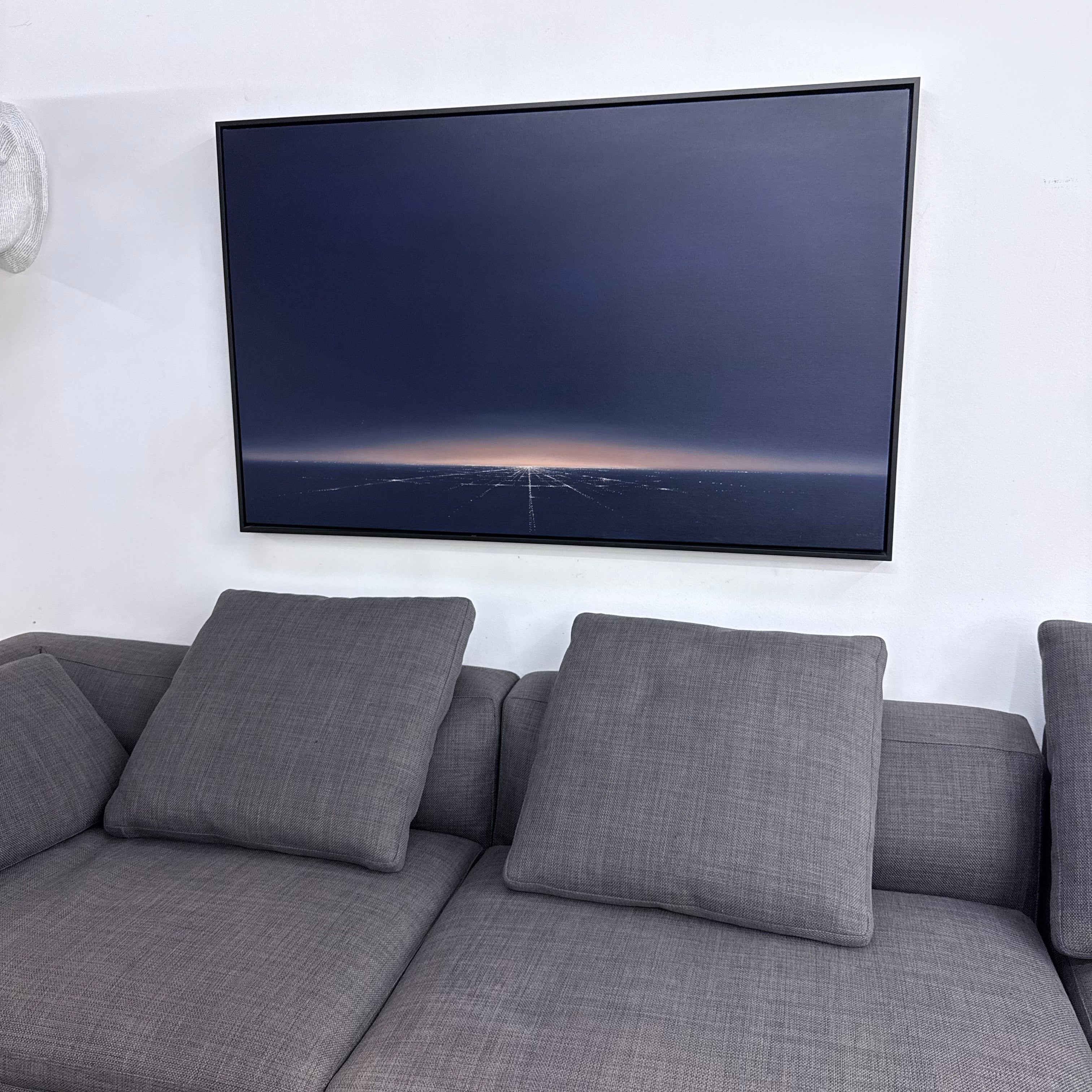 Inspired by "Painting: Western Arrival by Eric Nash" from Artist, this artwork shows a cityscape at dusk or dawn with a grid of lights and an orange glow on the horizon beneath a dark sky.