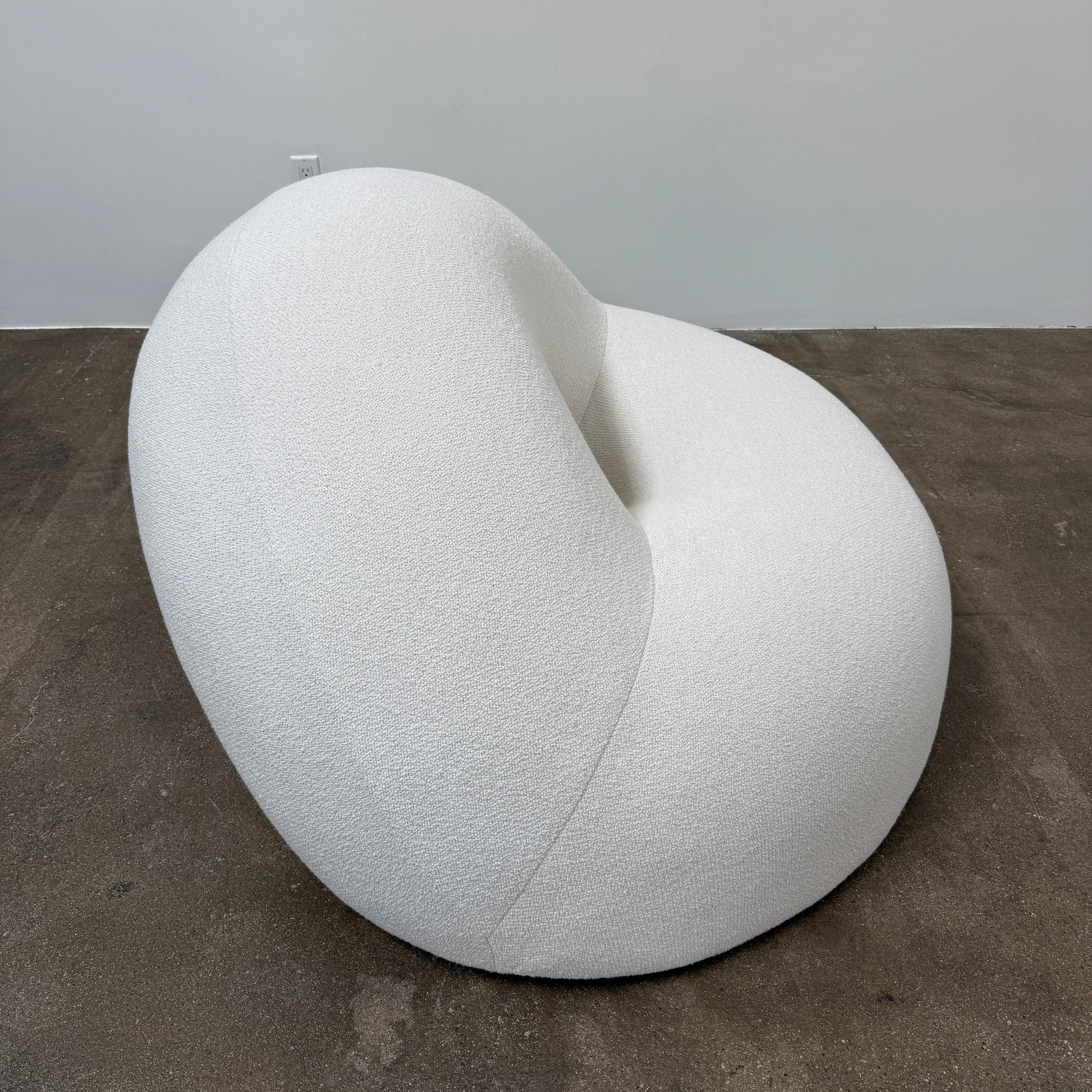 The Objects of Common Interest Tube Chair by Objects of Common Interest is a modern, light grey lounge chair with a donut-shaped design, rounded form, and central indentation, shown against a plain white background.