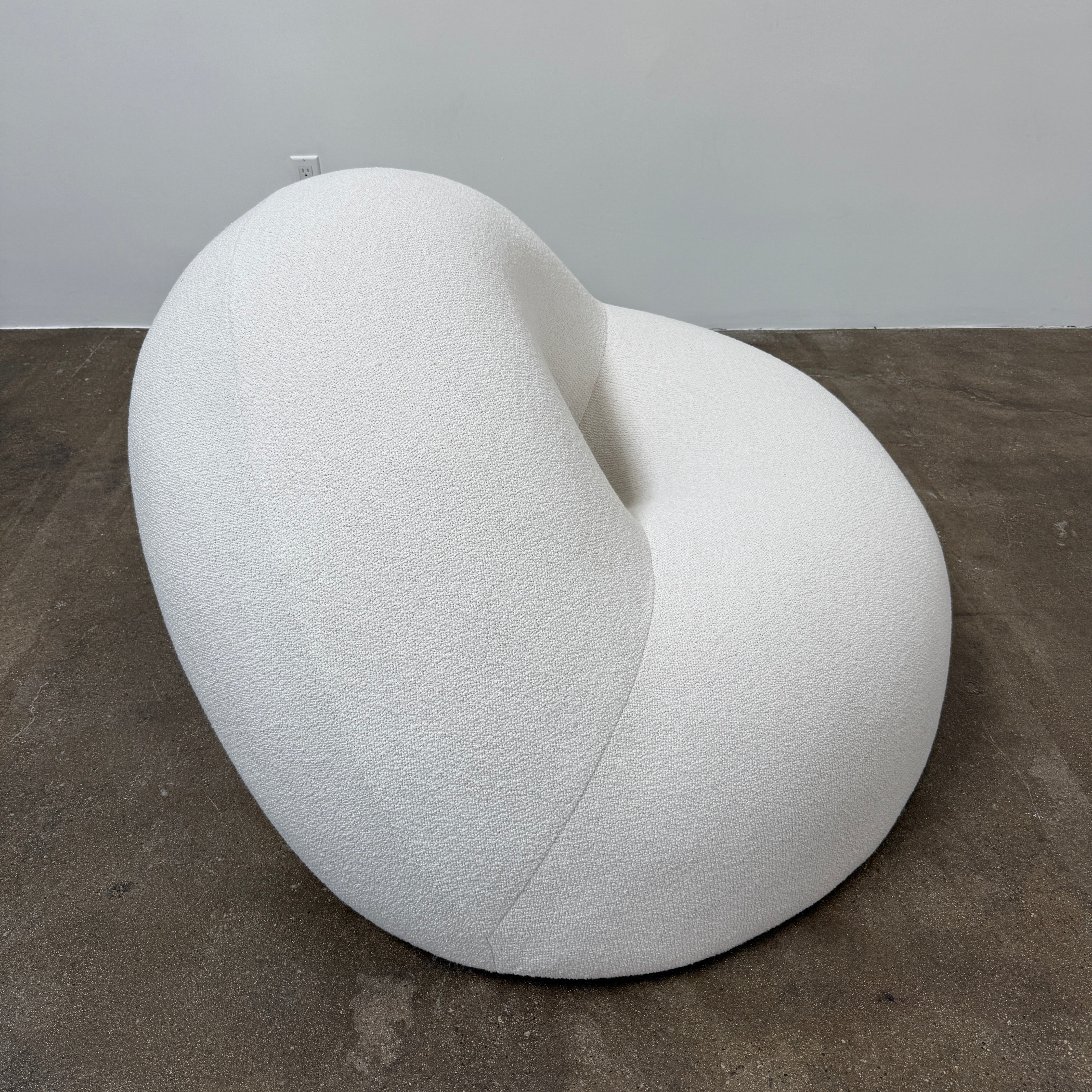 The Objects of Common Interest Tube Chair by Objects of Common Interest is a modern, light grey lounge chair with a donut-shaped design, rounded form, and central indentation, shown against a plain white background.