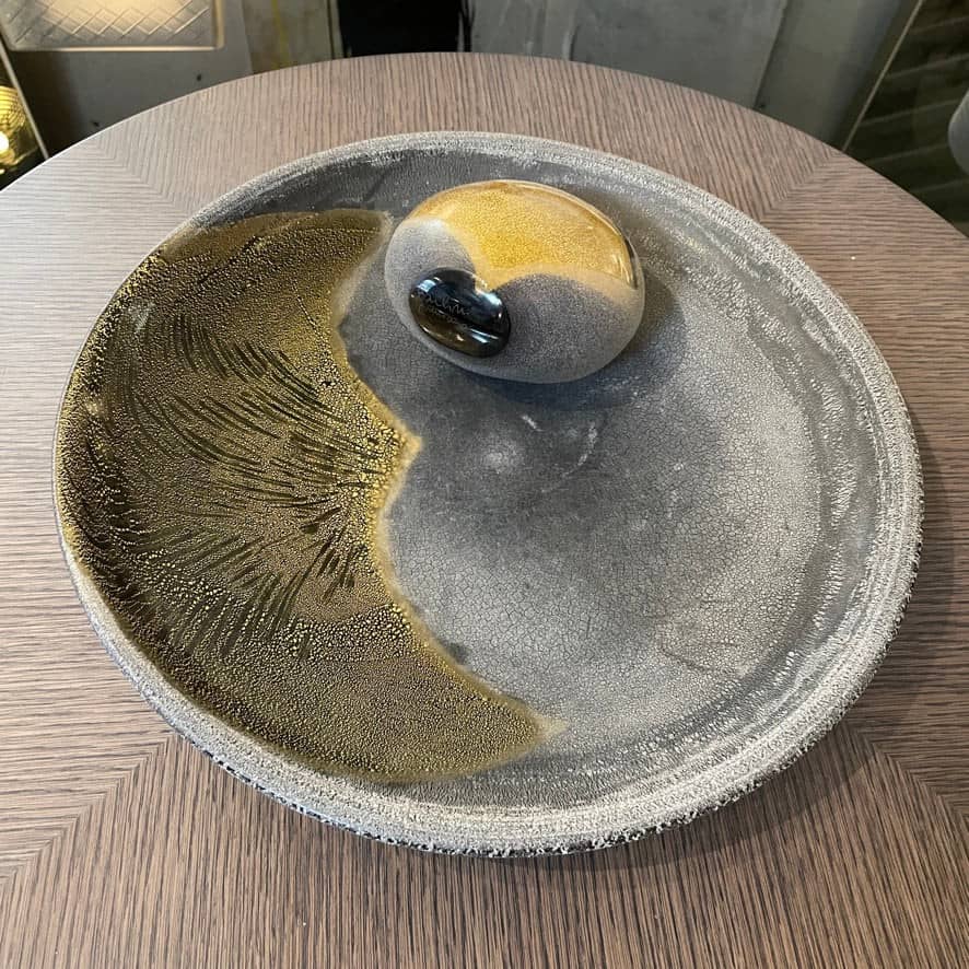 A round ceramic plate featuring a central raised circular design, predominantly gray with earthy bronze tones and textured streaks on one side, reminiscent of the artistry seen in the Alfredo Barbini Murano Glass Plate And Object.